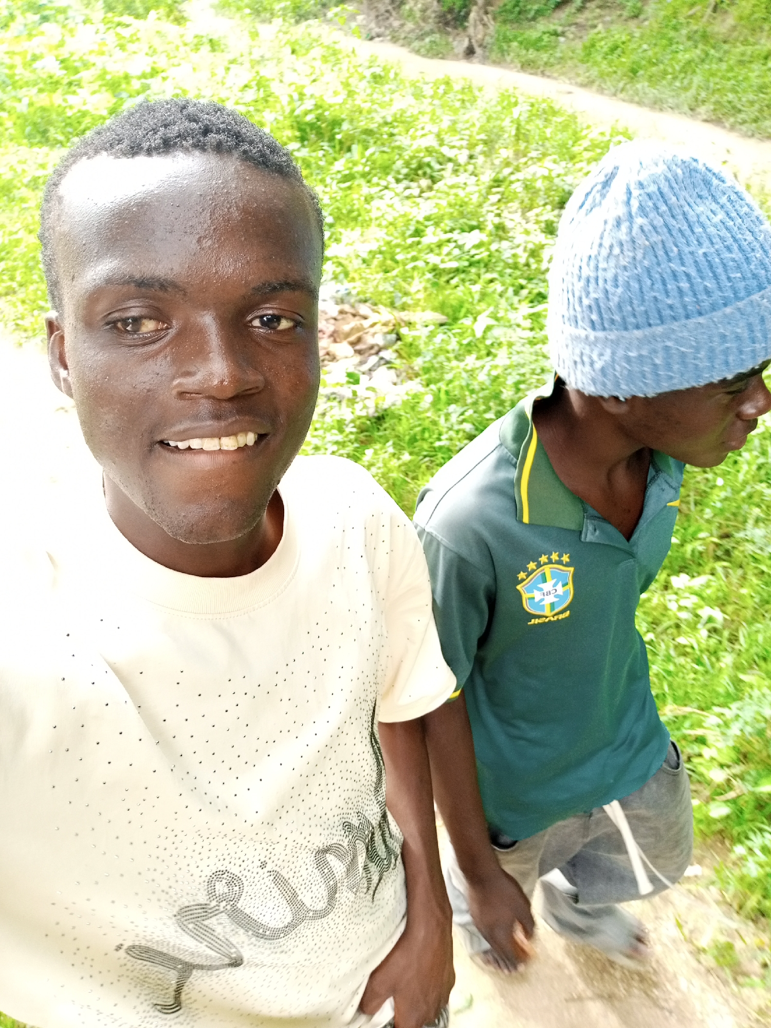 Two individuals are depicted in a close-up, eye-level, outdoor shot. The person in the foreground, facing the camera, has dark skin, short, textured dark hair, and is smiling, revealing their teeth. They are wearing a cream-colored t-shirt adorned with numerous small, dark dots and a script-like design across the chest. To the right, another individual, facing away from the camera, is wearing a light blue knit cap and a dark green polo shirt with a yellow collar. The polo shirt features a crest with stars and what appears to be the Brazilian flag. This individual is also wearing grey pants. The background consists of lush green foliage and a dirt path, suggesting a natural or semi-rural setting. The lighting indicates it is daytime, with bright ambient light. The image is taken from a selfie perspective, with the camera held at a slightly high angle.