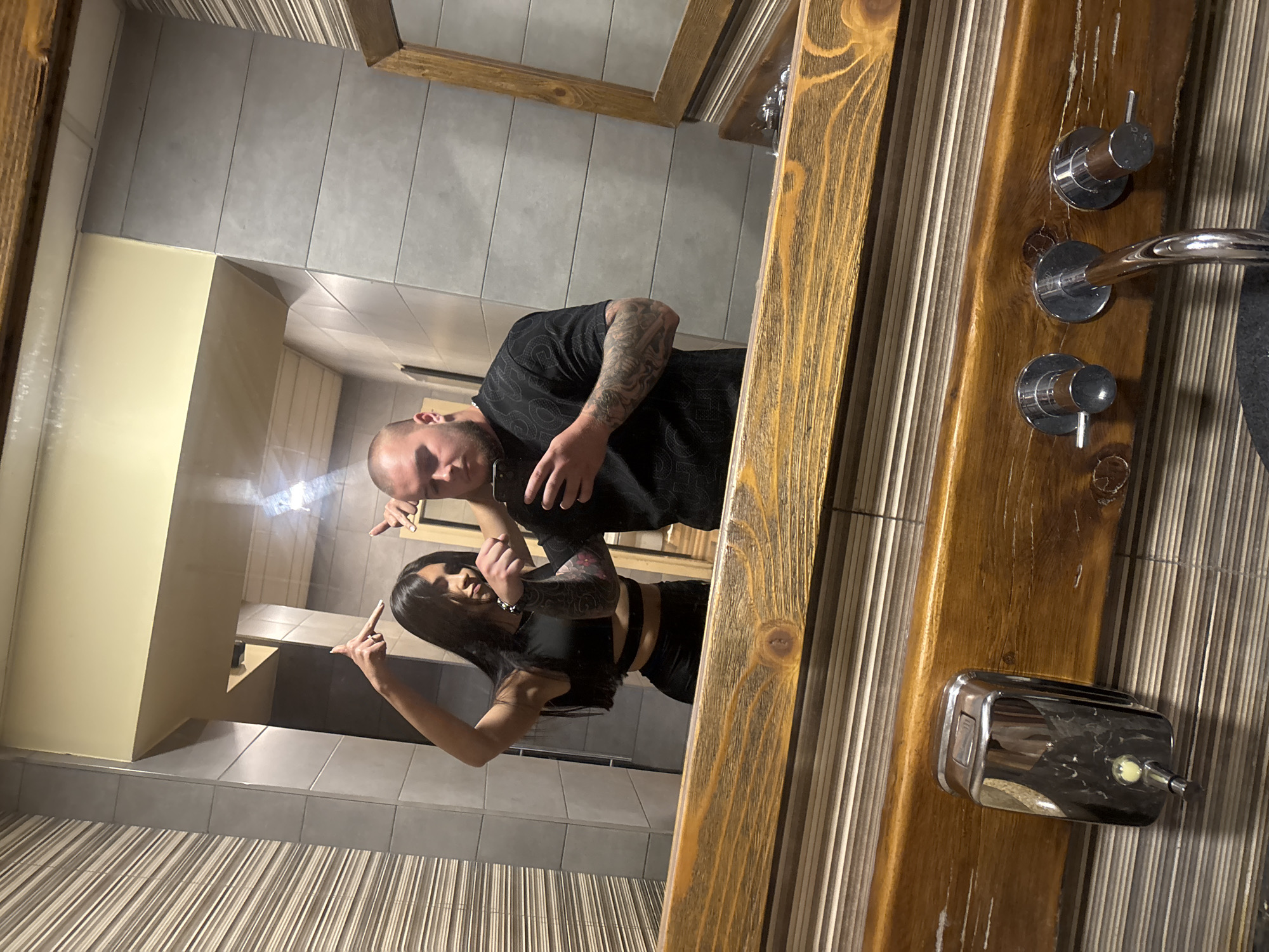 A bald man and a woman with long dark hair are reflected in a mirror. They are both tattooed and wearing dark clothing. The man is holding a mobile phone. The mirror has a rustic wood frame. The location appears to be a bathroom or restroom. The scene includes a sink, faucet handles, and a soap dispenser mounted on the wall. The city of Pleven, Bulgaria is not discernible from the image.