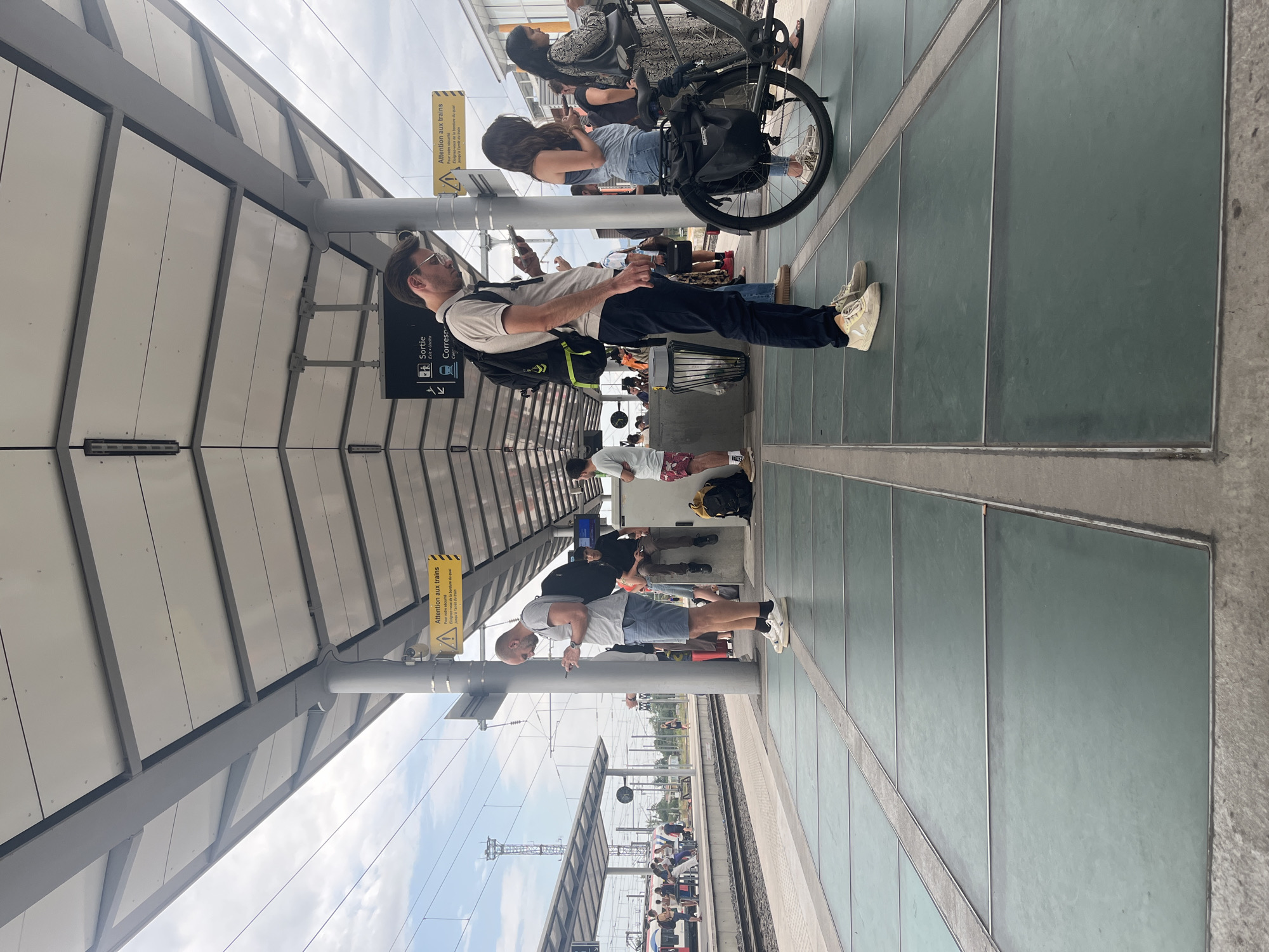 The image depicts a street view in Annemasse, France, specifically a train station platform. Several individuals are visible, mostly standing and engaged with their mobile phones. A man in the foreground is wearing a light-colored polo shirt, dark pants, and white sneakers, with a backpack on. To his right, a woman in a blue tank top and jeans stands next to a bicycle. Other people are scattered on the platform, some near a train in the background. The platform features a modern design with a translucent tiled surface and a covered canopy overhead supported by pillars. Yellow warning signs with black text, indicating "Attention aux trains" (Attention to trains), are prominently displayed on the pillars. The environment suggests a busy public transportation hub under a cloudy sky.