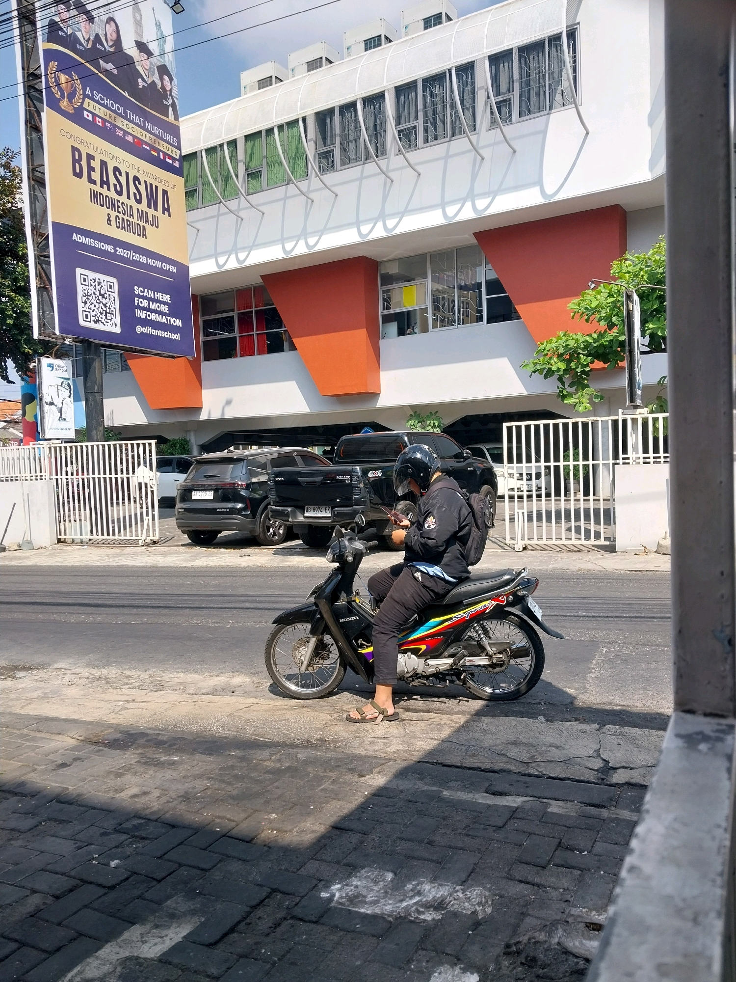 In Depok, Indonesia, a person wearing a black jacket, black pants, a black helmet, and sandals is sitting on a black motorcycle. The motorcycle has colorful decals on its side. The individual is looking down at a mobile phone held in their hands. The motorcycle is parked on a paved surface next to a street. In the background, a modern white building with orange architectural accents is visible. A large banner hangs on the left side of the building, advertising a school and a scholarship opportunity, with the text "BEASISWA INDONESIA MAJU & GARUDA" and "ADMISSIONS 2027/2028 NOW OPEN." Several cars, including a black SUV and a black pickup truck, are parked behind a white fence in front of the building. Green trees and a lamppost are also present in the environment. The sky is clear and the scene appears to be illuminated by bright sunlight, casting shadows on the ground.
