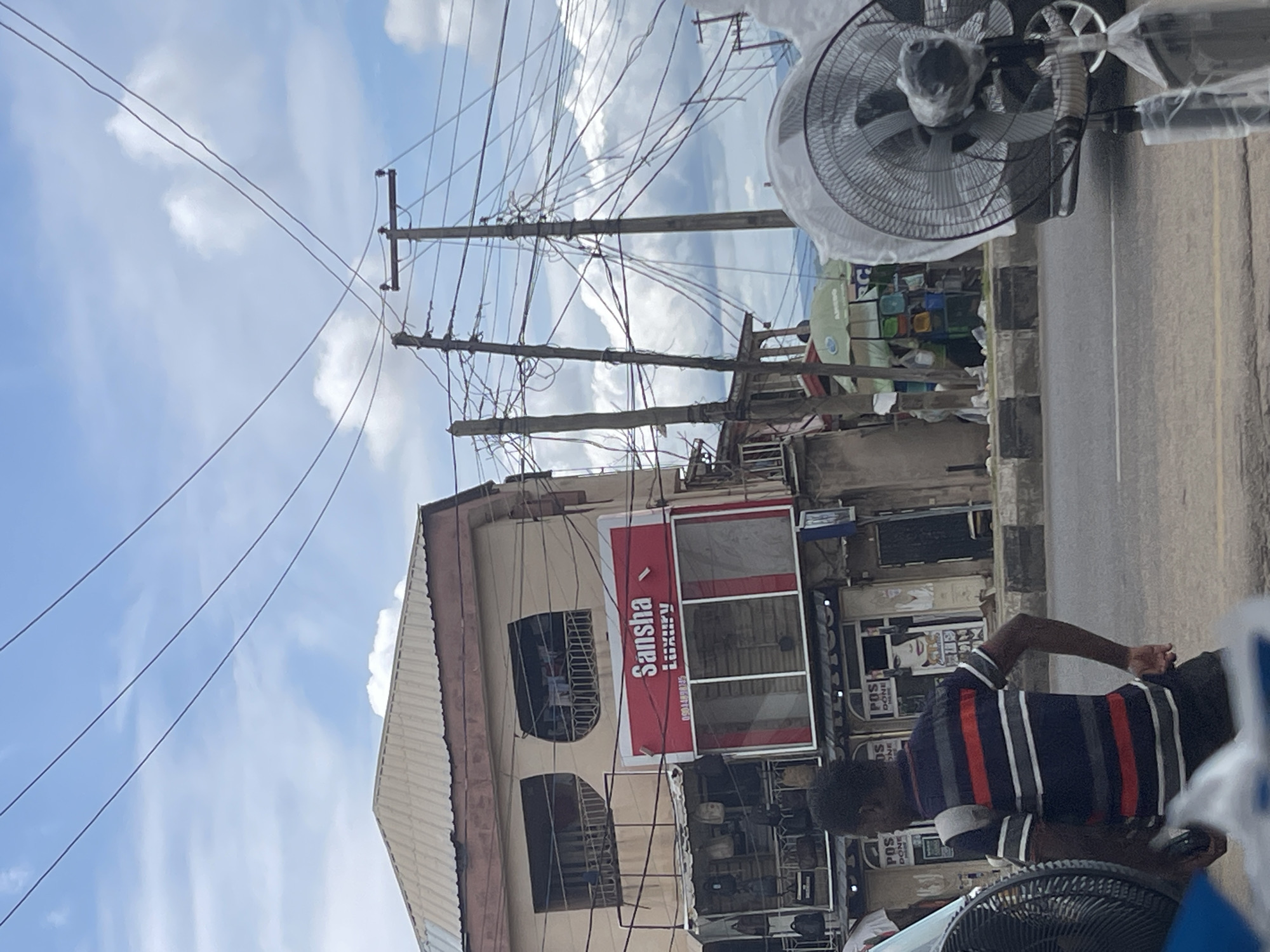 A street scene in an urban environment under a cloudy sky features a multi-story building with a red sign that reads "Sansha Luxury" in white lettering. Below this sign, shops are visible with displays of merchandise. A man wearing a horizontally striped shirt walks away from the camera, carrying a backpack. In the foreground and to the right, a large electric fan, partially covered in plastic, stands on the sidewalk. Utility poles with a dense network of electrical wires crisscross the sky behind the building. The scene appears to be a busy commercial area with signs of everyday life and commerce.