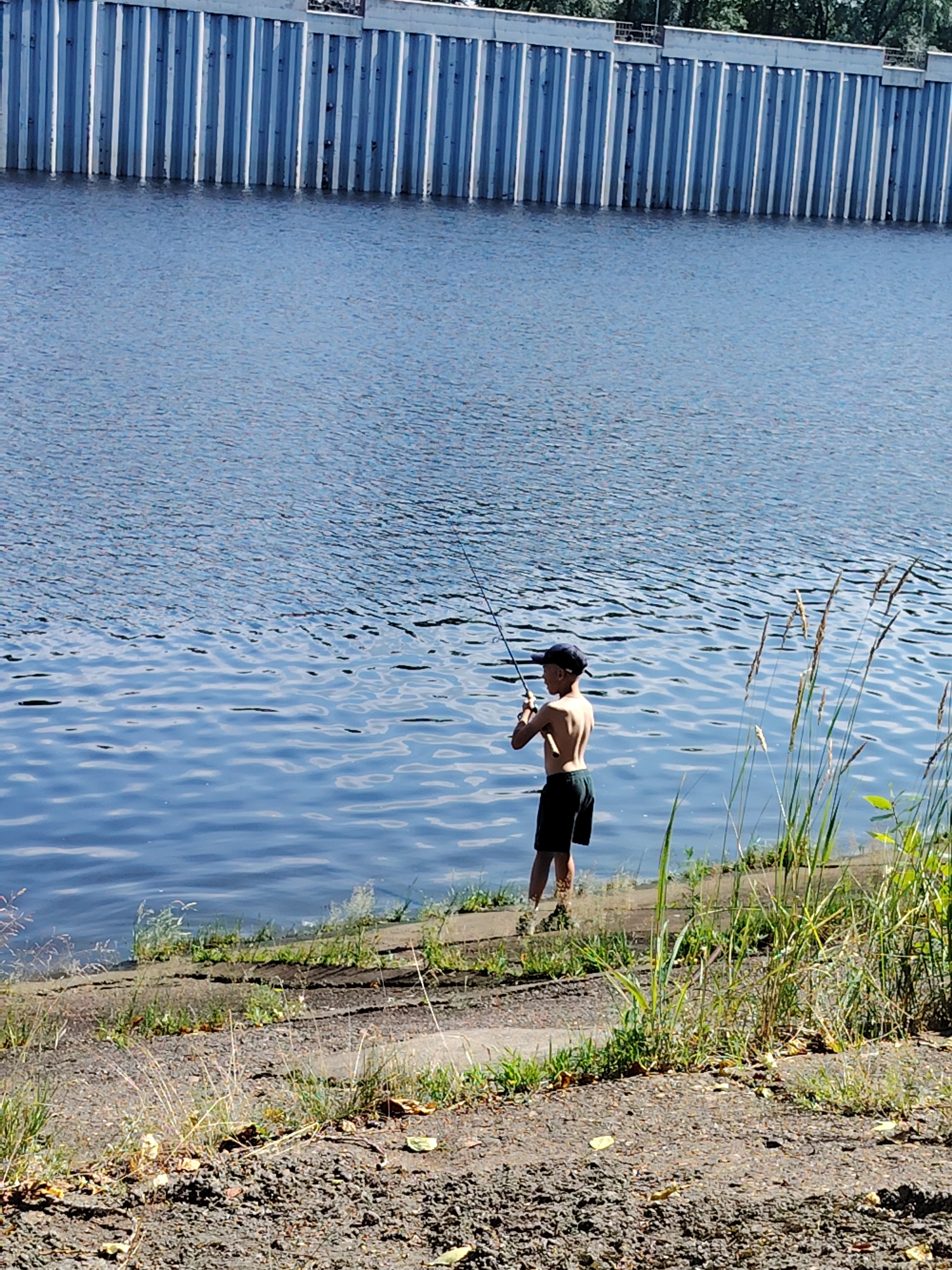 The image depicts a young boy fishing on the bank of a river in Chaikovsky, Russia. The boy, wearing a dark baseball cap and dark shorts, stands shirtless in the shallow water, holding a fishing rod. The river's surface is rippled, reflecting the bright sunlight. In the background, a tall, grey, ribbed wall, likely a dam or retaining structure, lines the opposite bank. The foreground shows a textured shoreline composed of gravel and sparse vegetation, including dry grasses and some green shoots. The scene is captured on a clear, sunny day.