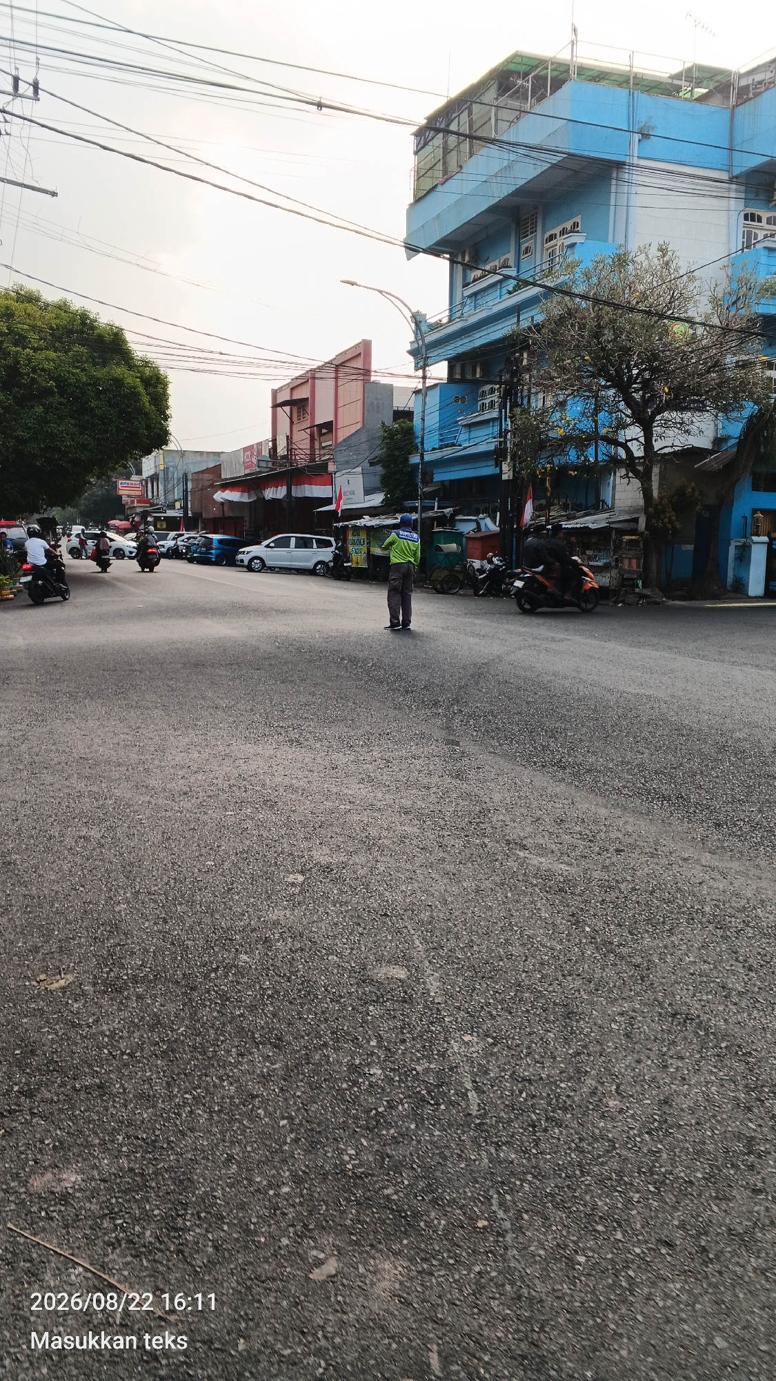 A wide, asphalt road in what appears to be a commercial area in Malang, Indonesia, is captured from a low perspective. The sky is overcast and bright. On the right side of the street, a multi-story blue building with numerous windows stands prominently. Next to it, a tree with sparse yellow fruit is visible. A person wearing a green shirt and dark pants stands in the middle of the road, facing left. Several parked cars and motorcycles line the street on both sides, with some vehicles in motion. Buildings with red and white Indonesian flags are visible further down the road. A large tree with green foliage is on the left side of the image. The timestamp "2026/08/22 16:11" and the text "Masukkan teks" are present in the bottom left corner.