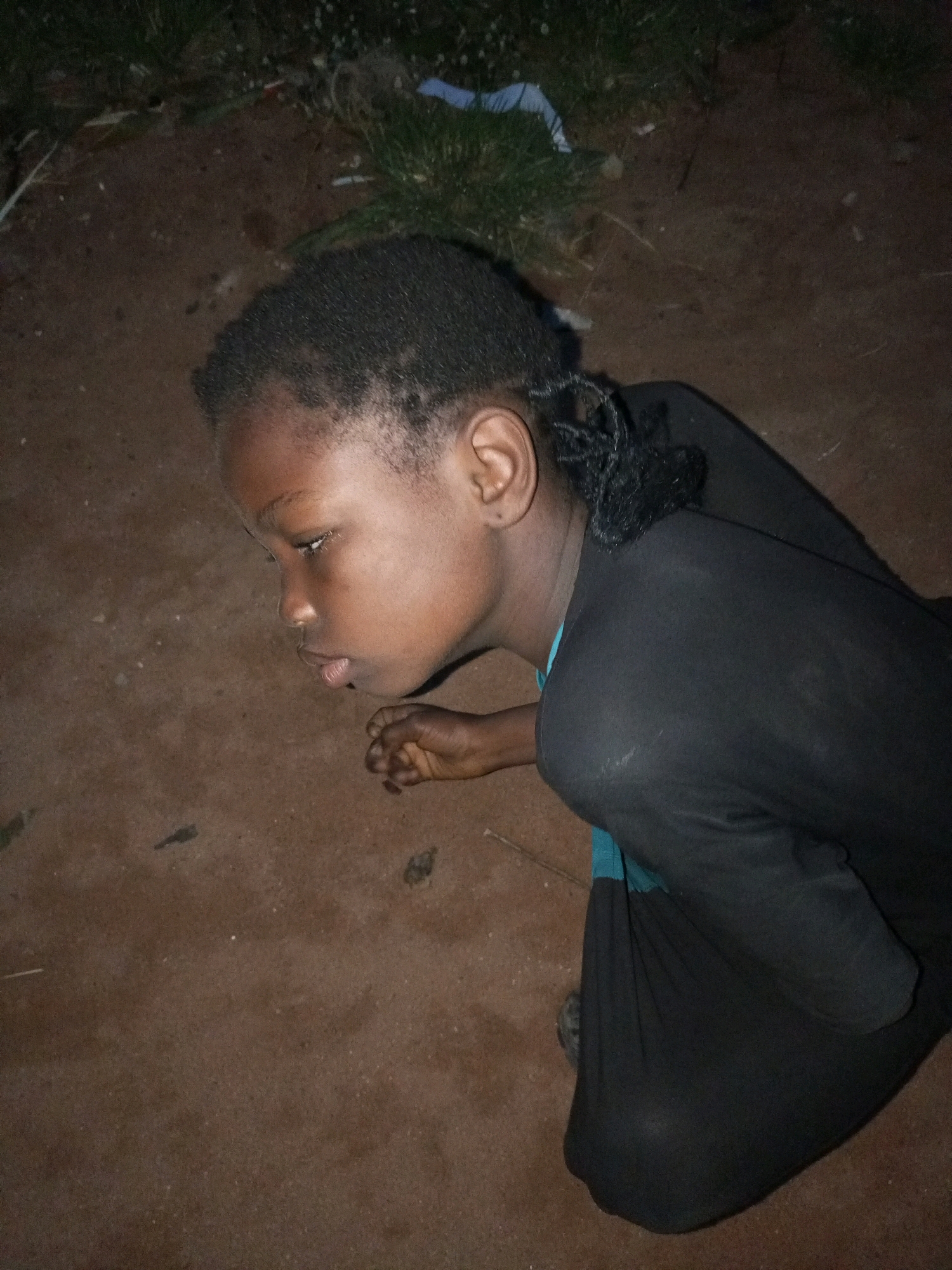 A young Black female is kneeling on the ground outdoors. She is wearing a black garment. Her hair is styled in braids. The ground is composed of dark brown soil and some sparse green vegetation is visible in the background. The scene appears to be illuminated by artificial light. The location is identified as Tempure, Nigeria.