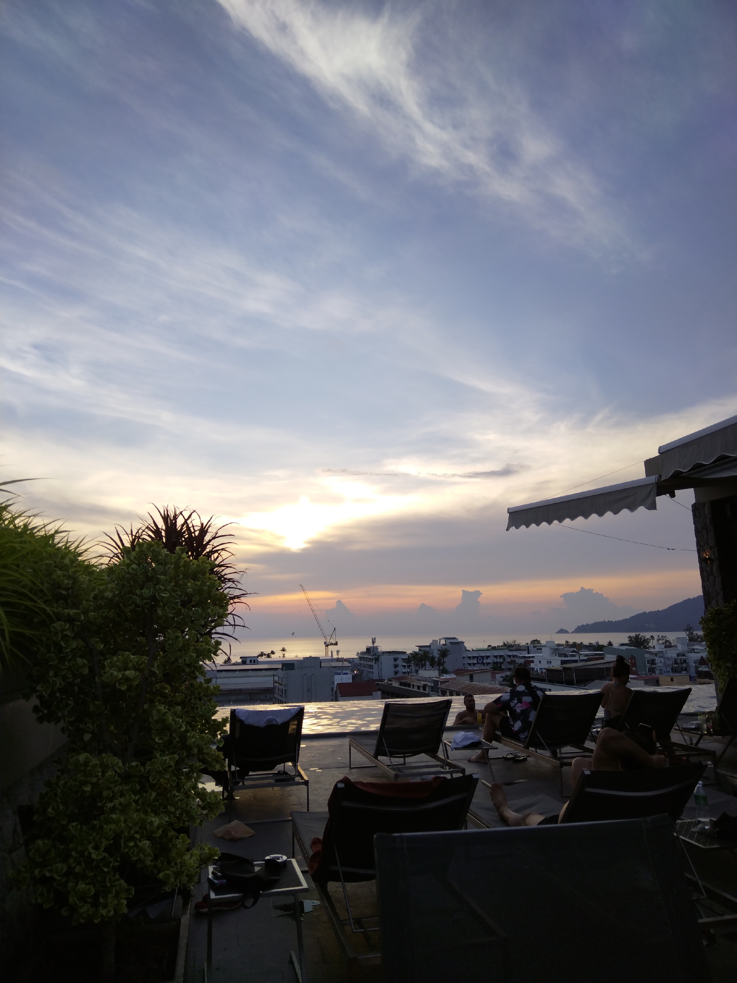 The image depicts a rooftop view of Patong, Thailand, at sunset. In the foreground, several empty lounge chairs are arranged on a dark-tiled surface. To the left, a lush green plant with spiky leaves is prominent. In the middle ground, a few people are relaxing on lounge chairs, some appearing to be engaged in conversation or looking at their phones. Beyond the rooftop, a cityscape of buildings stretches towards the ocean, with a few cranes visible in the distance. The sky is a gradient of soft blues, oranges, and yellows, indicative of the setting sun. The overall scene suggests a tranquil evening at a rooftop establishment overlooking the city and the sea.