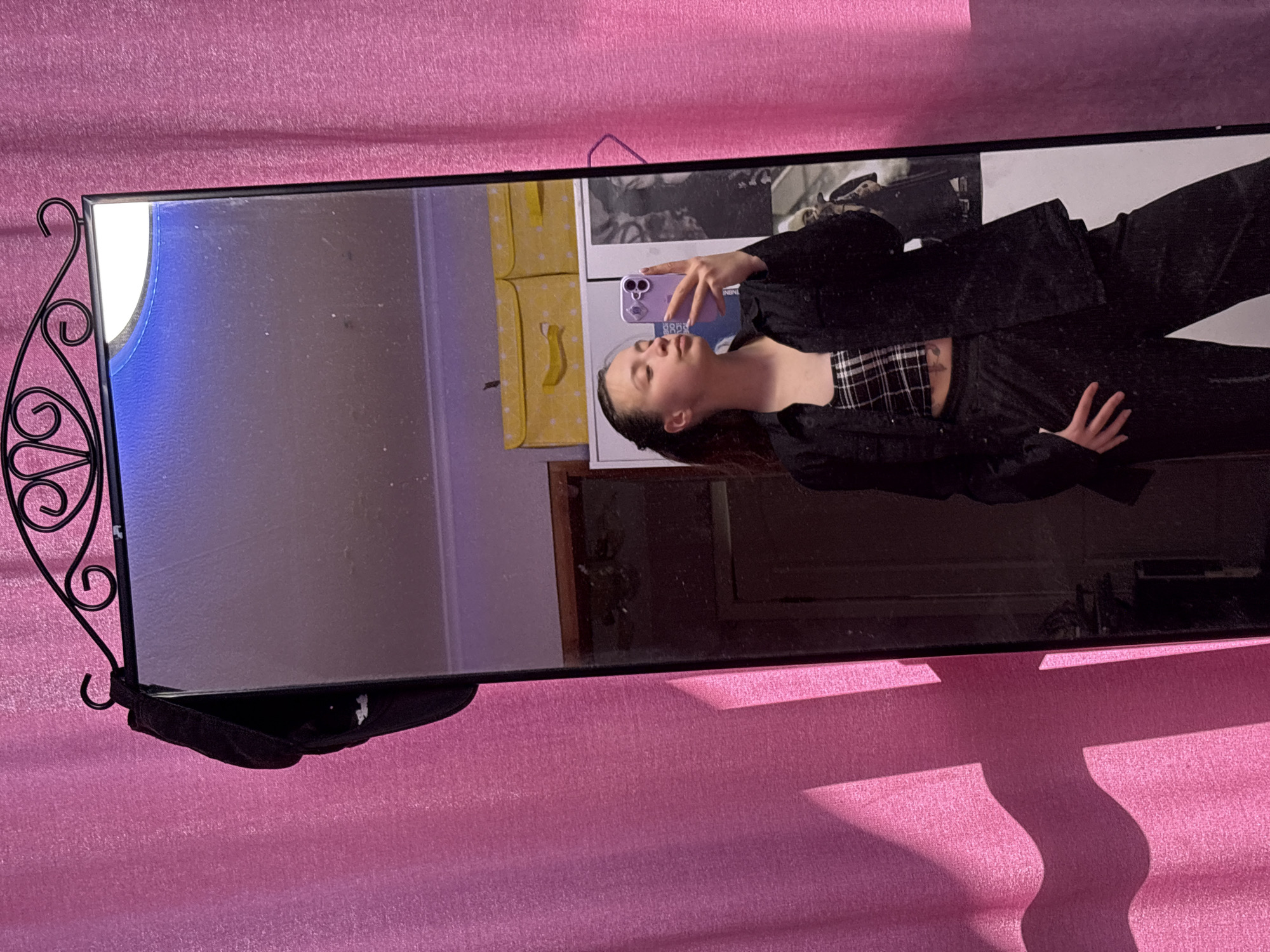 A horizontally oriented photograph captures a reflection in a framed mirror. The mirror's frame is black with decorative wrought iron elements on its left side. Reflected in the mirror is a woman holding up a purple smartphone, apparently taking a selfie. She is wearing a plaid top, a black jacket, and black pants. Behind her is a wall with a yellow storage container and a black and white picture hanging on the wall. The mirror is positioned against a pink textured surface. The location is Прикамский, Russia.