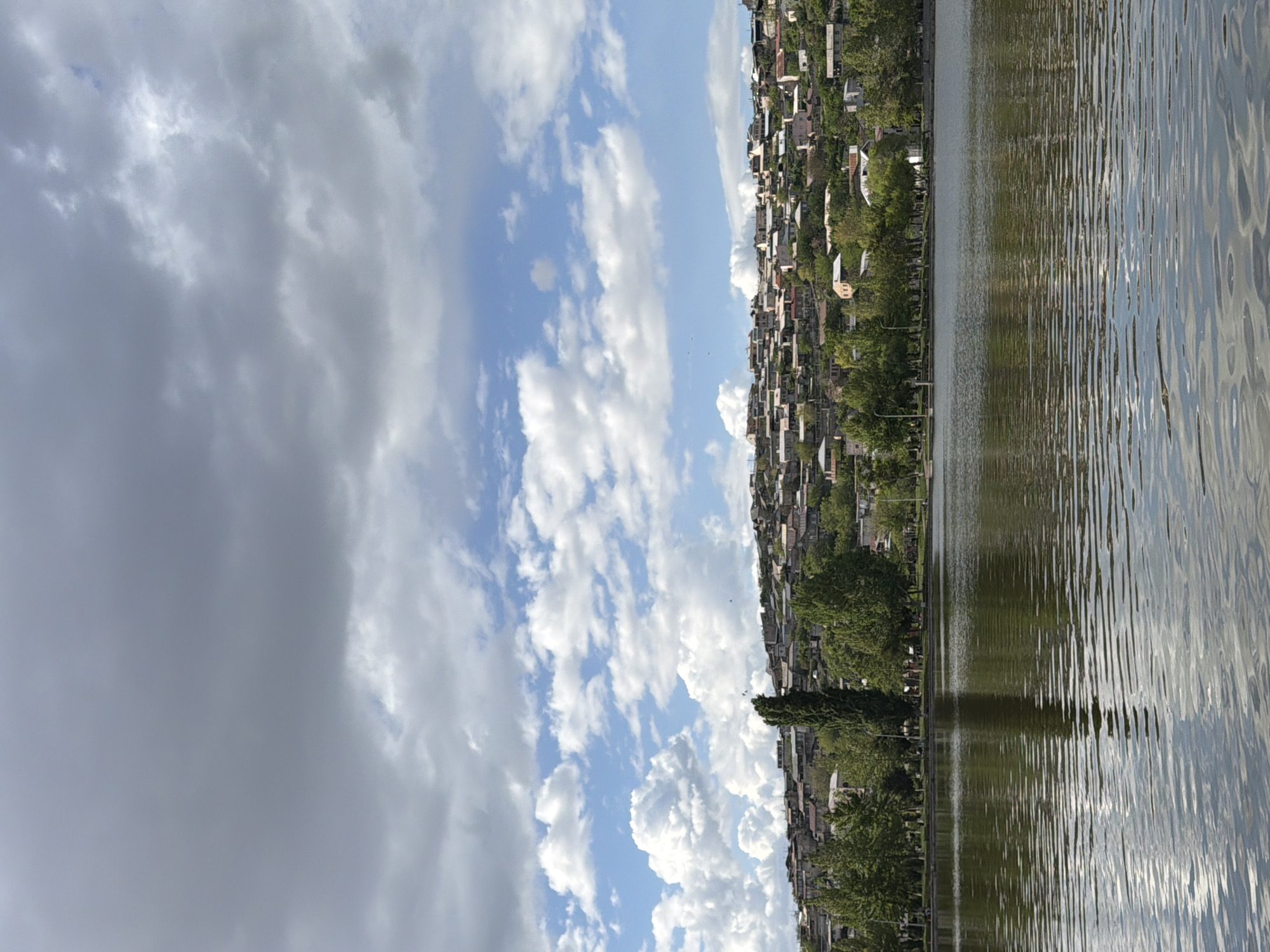 The image depicts a view of Yerevan Lake, Armenia, alongside a cityscape under a cloudy sky. The lake surface exhibits rippling reflections. The cityscape consists of closely packed buildings with interspersed greenery. The sky is filled with a combination of dark, heavy clouds and patches of blue sky dotted with fluffy white clouds.
