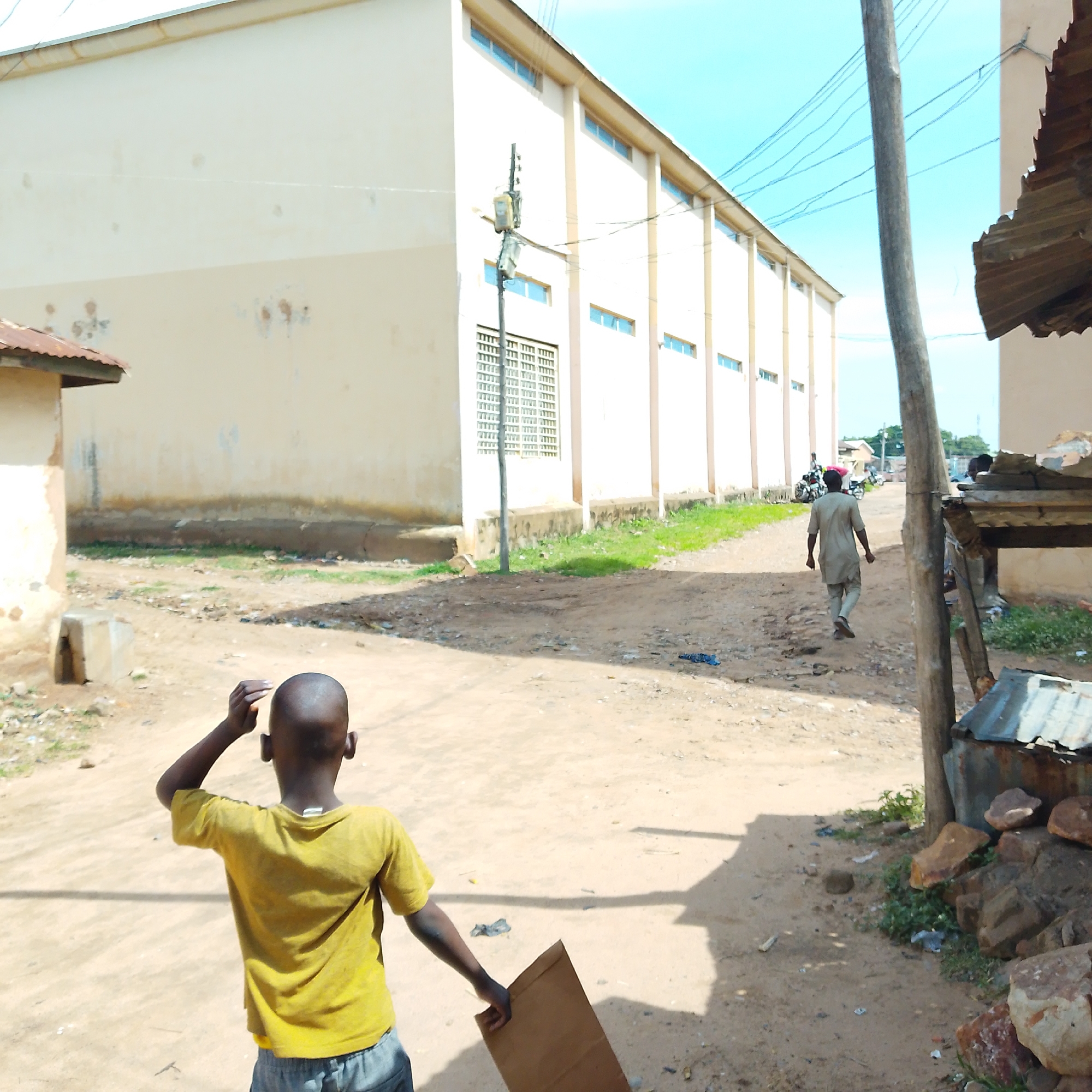 This image depicts an outdoor scene in Pantame, Nigeria, likely in an urban or semi-urban area. In the foreground, a young boy with a shaved head, wearing a yellow t-shirt and grey shorts, stands with his back to the camera, holding a piece of cardboard. He is facing away from the viewer, towards a light-colored, elongated building with multiple windows. To the right of the boy and further down the dirt path, an adult male, dressed in a light-colored tunic and trousers, walks away from the camera. He appears to be carrying something on his head, possibly covered with a pink cloth. Behind him, a motorcycle is visible with two individuals on it. The environment is characterized by a dirt or sand-covered ground, with scattered debris and some patches of grass and vegetation. A wooden pole with electrical wires runs vertically through the scene, and a corrugated metal structure is partially visible on the right side of the image. The sky is bright blue with some wispy clouds. The large building in the background has a light-colored facade and rows of windows with grey frames.