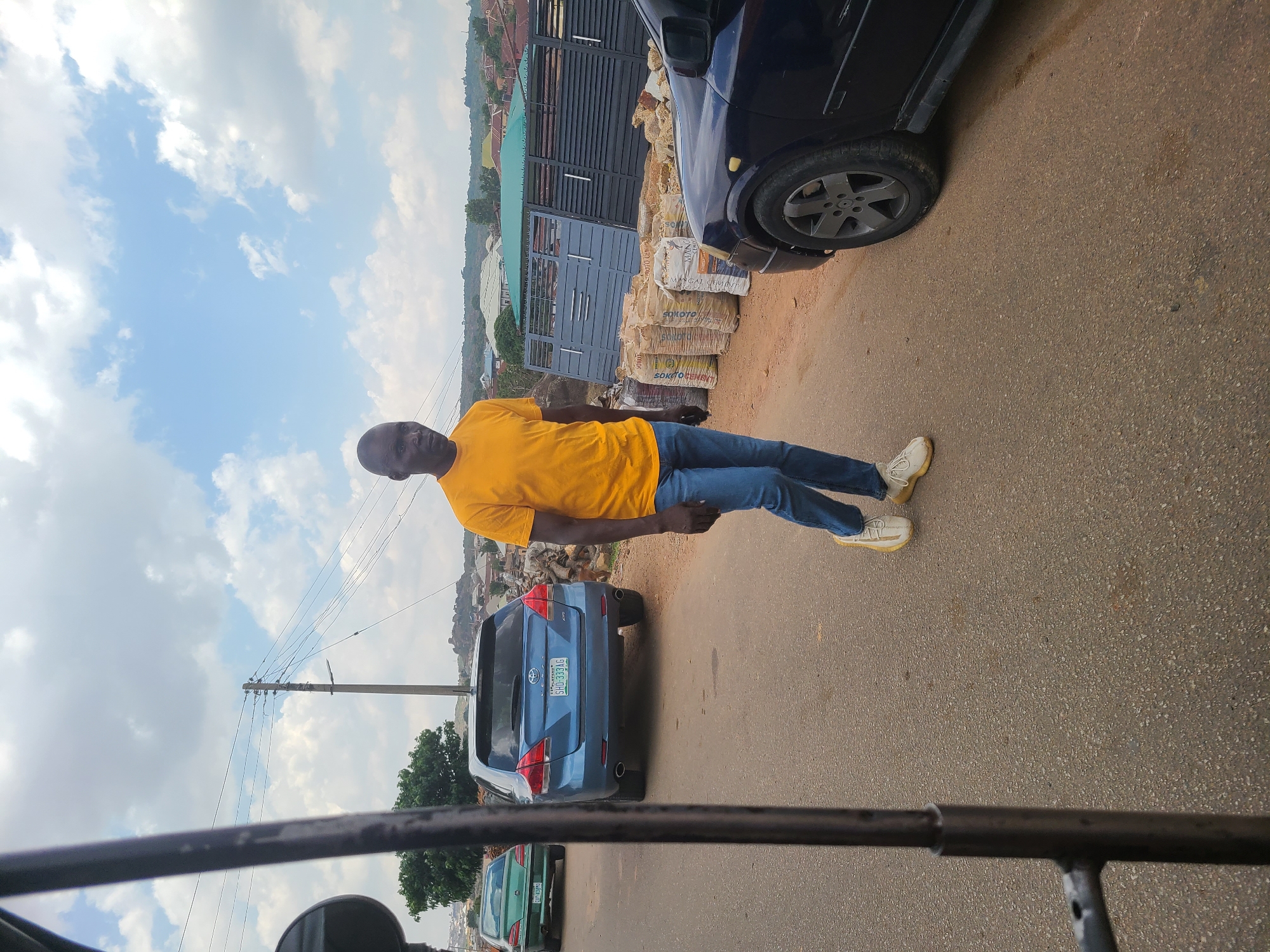 A man wearing a yellow shirt and blue jeans stands on the side of a road in what appears to be a residential area in Jos, Nigeria. Bags of construction materials are stacked near a dark-colored vehicle. There is a blue SUV parked behind him and a green car is visible further down the road. Utility poles with wires run along the side of the road. The sky is cloudy. The image seems to be taken from inside a vehicle.