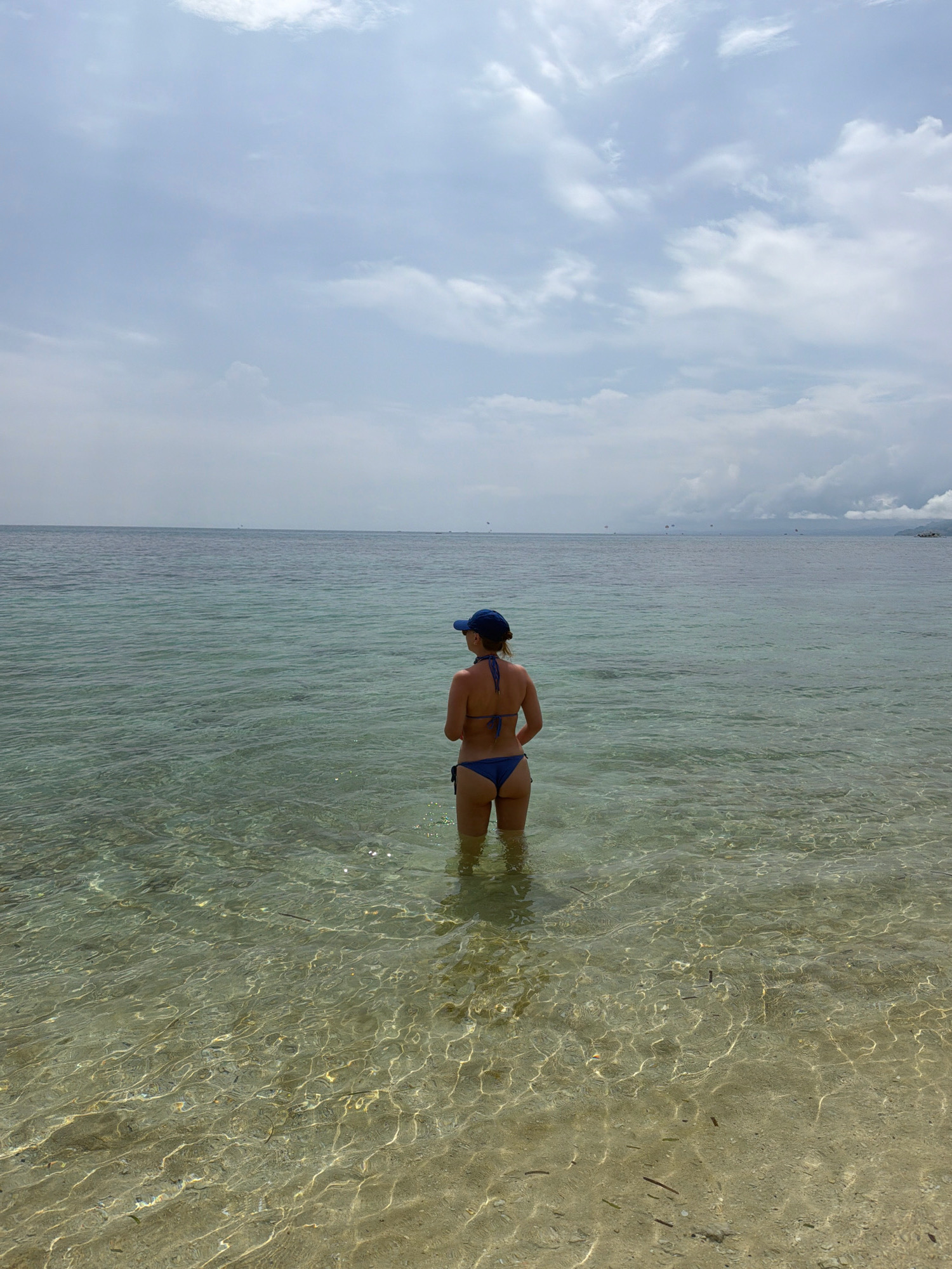 In the city of Malay, Philippines, a woman stands in waist-deep, clear turquoise water on a beach. She is facing away from the camera, wearing a blue bikini and a blue baseball cap. The water is shallow and the sandy seabed is visible through the clear water, with some small pieces of debris scattered on it. The sky above is overcast with light gray clouds. In the distance, there is a faint horizon line separating the sea and sky, with a few indistinct shapes that could be boats or other structures. The overall scene depicts a calm, tropical beach setting.