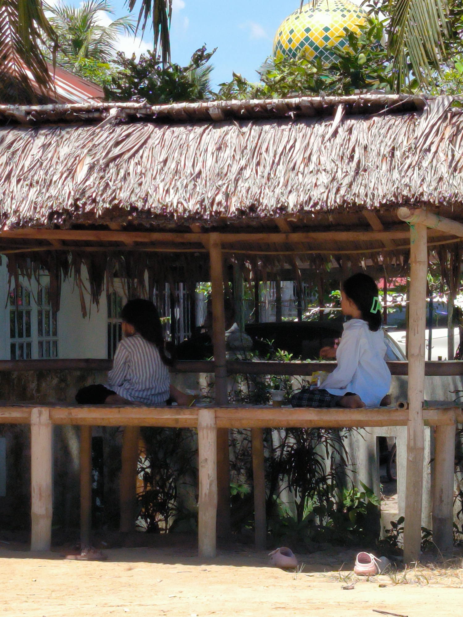 The image shows two young girls sitting on a wooden structure with a thatched roof. They appear to be in a rural or semi-rural setting, possibly in the city of Merbopuntong, Indonesia.
The girl on the left is wearing a striped shirt and dark-colored pants. The girl on the right is wearing a light-colored shirt and plaid bottoms, with a green barrette in her hair. Both are barefoot.
The structure they are sitting on is made of light-colored wood. Beneath the structure, on the ground, are a pair of pink sandals.
In the background, there is a house with white doors and windows. There are also various plants and trees visible, including palm trees. In the upper right corner, a building with a yellow and green domed roof can be seen. The sky is blue with scattered clouds. The ground appears sandy.
