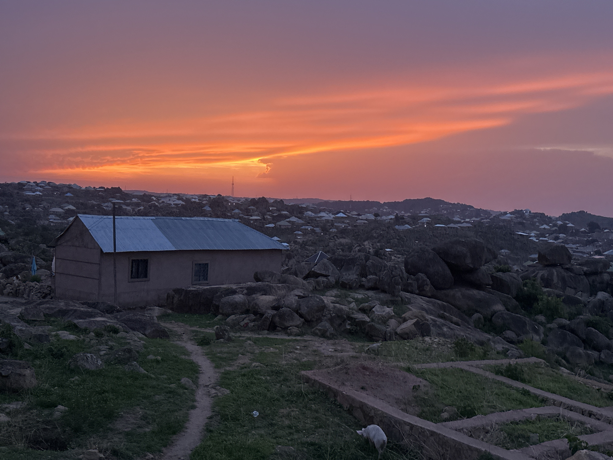 A wide-angle shot captures a dwelling in Jos, Nigeria, set against a landscape of granite formations and distant buildings under a twilight sky.
The small, rectangular building has a tin roof, pale earthen walls, and two windows. A dirt path leads toward the house, which sits among large rocks.
In the background, the city's architecture blends with the natural terrain of rounded, dark granite boulders. The skyline displays an orange and purple gradient.
A white animal is in the lower right corner of the image. In front of it there are rectangular planters.