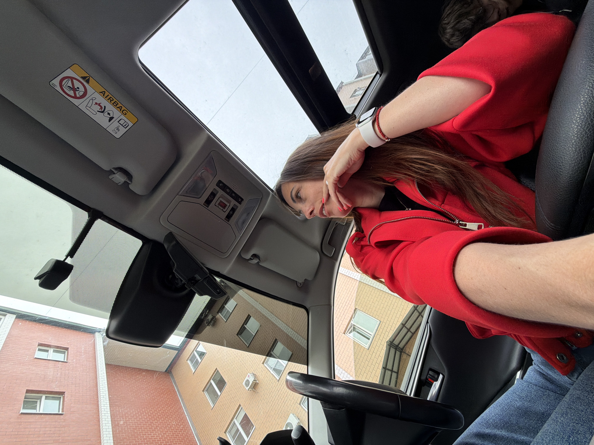 A woman in a red jacket sits in the driver's seat of a car with a sunroof. The car is parked on a street in Birobidzhan, Russia, as indicated by the surrounding architecture. The woman wears a white smartwatch on her left wrist. The car's rearview mirror is visible. Buildings are reflected on the windshield.