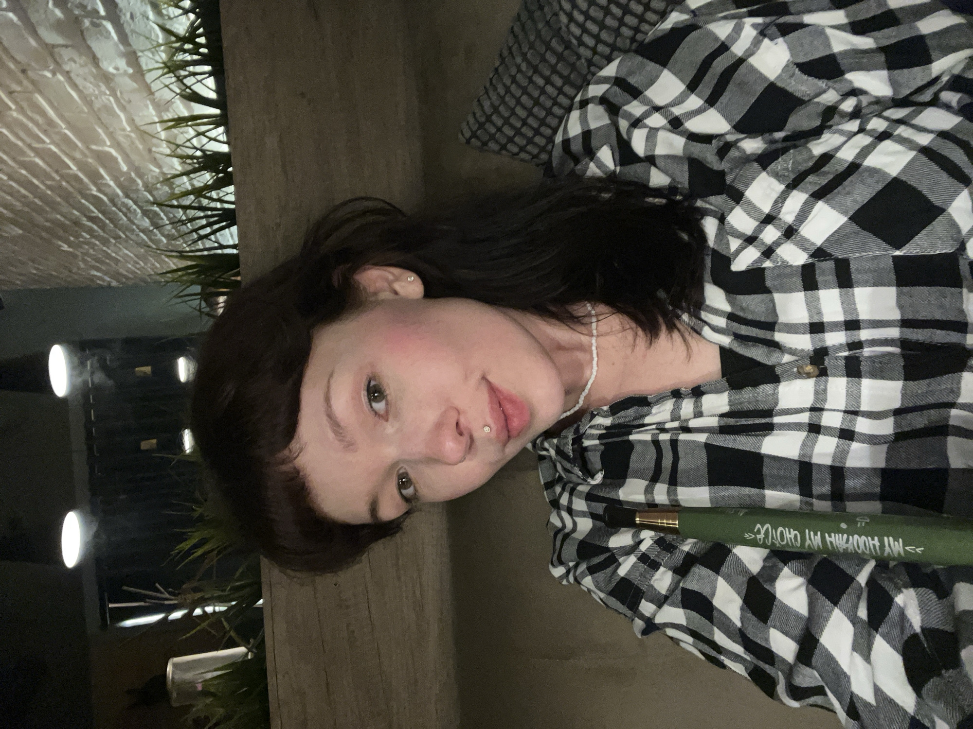 A young woman is photographed outdoors in a location that has not been confirmed to be in Chaikovsky, Russia. She has dark brown hair with bangs, fair skin, and a lip piercing. She is wearing a black and white checkered flannel shirt and a beaded necklace. She is seated or lying down on a wooden surface or bench. A green object with white writing is partially visible in the lower right corner, but its identity and content are unclear. Behind her, there is a brick wall with plants growing along it. There are also lights visible in the background. The image has a slightly rotated orientation.