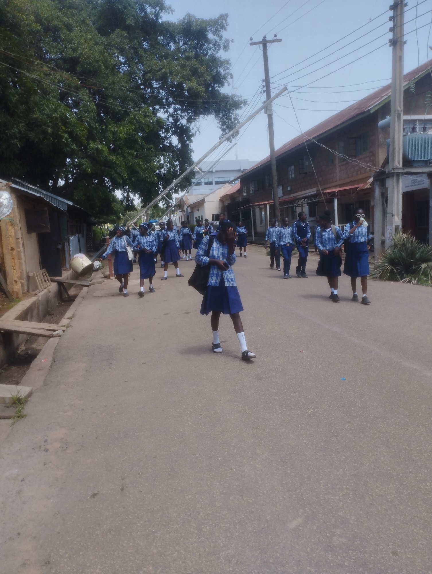 A group of young people, likely students in uniform, are walking down a paved street in a city. The uniforms consist of blue and white checkered shirts and blue skirts or shorts, with some wearing white socks and dark shoes. They are walking in the same direction, some in pairs and others individually. In the background, there are buildings with varying architectural styles, some appearing older with corrugated metal roofs, and others more modern. Utility poles with numerous wires are visible along the street. Lush green trees are present on the left side of the image, and some vegetation can be seen around the buildings. The overall impression is of an urban setting with residential or commercial structures and a clear sky. The presence of students in uniform suggests a school or educational area. The scene appears to be in Jos, Nigeria, given the context provided.