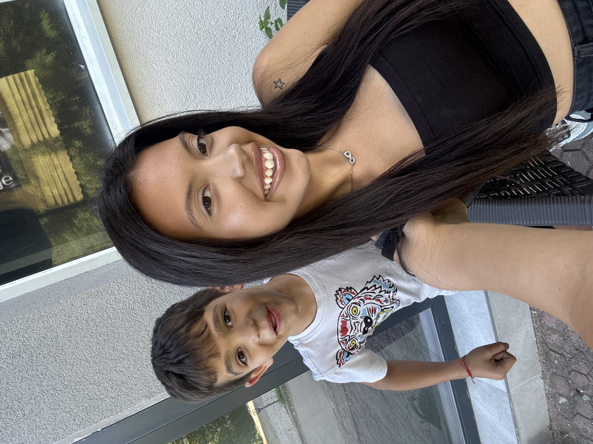 This is a selfie taken by a young woman with long, dark hair. She is wearing a black crop top and jeans. She is smiling broadly and has a necklace with an infinity symbol. To her left and slightly behind her is a young boy with short, dark hair. He is wearing a white t-shirt with a colorful tiger graphic on it and a red string bracelet on his left wrist. The background appears to be an outdoor setting, possibly a patio or garden, with a textured wall and a window. The overall impression is a candid and happy moment captured between two individuals. The context suggests they might be in a residential area or a cafe in Sofia, Bulgaria, given the provided location information.