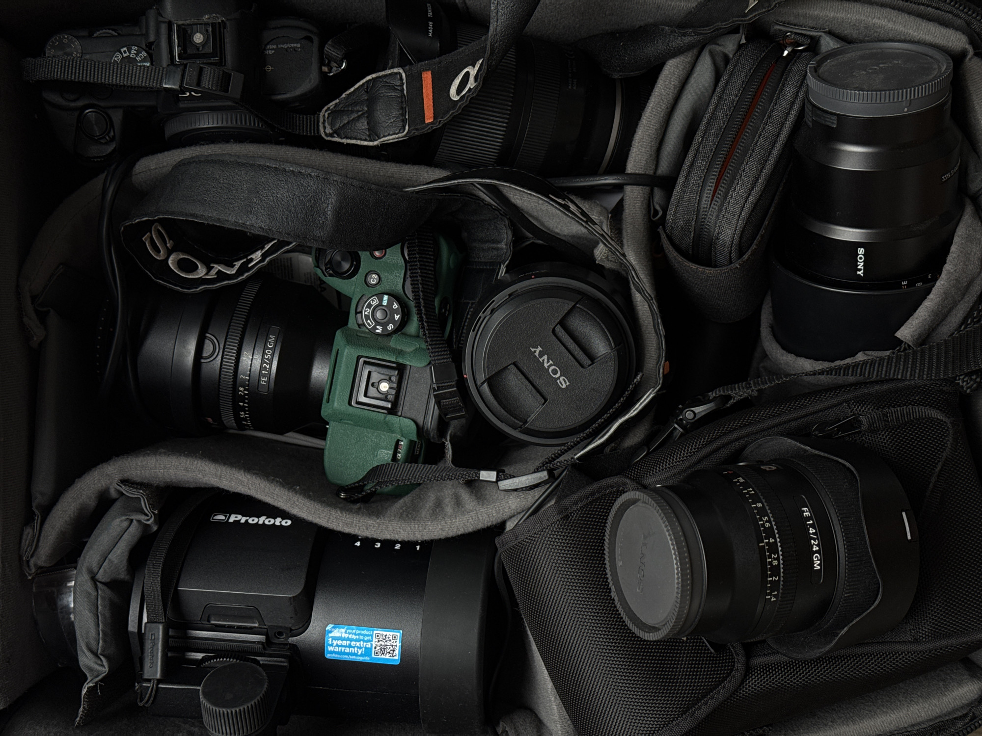 This top-down, high-angle shot captures a professional array of camera equipment neatly organized within a padded camera bag, suggesting preparation for a shoot or organized storage in Dubai, United Arab Emirates. The collection includes multiple camera bodies and lenses, predominantly from Sony, along with a lighting unit from Profoto.
At the center, a dark green Sony Alpha camera body is conspicuous, mounted with a Sony FE 1.2/50 GM lens, featuring aperture markings like "1.2," "2," "2.8," and "4." Above it, another Sony Alpha camera body (black) displays a mode dial with settings such as "SCN," "P," "A," and "M," and bears a black strap with the white "α" logo. Another black Sony lens is visible in the upper right quadrant, marked "SONY" on its side, and another lens cap also says "SONY" with the text "31090" beneath it. A third Sony FE 1.4/24 GM lens, complete with aperture markings from "1.4" to "16," rests in the bottom right corner, covered with a "SONY" lens cap. A partial view of a Tamron lens, with text "HA046 TAMRON," is also discernible.
A large black Profoto lighting unit or power pack is situated in the bottom left, clearly displaying the "Profoto" logo and a bright blue sticker advertising "1 year extra warranty!" with a QR code and the URL "profoto.com/sets." A small, grey, zippered accessory pouch is tucked into a compartment next to a large lens. The overall aesthetic is dark and professional, highlighting the high-end photographic gear.