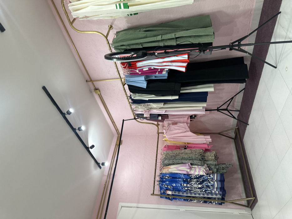The image depicts the interior of a clothing boutique in Mexico City, Mexico. The room features textured pink walls and a white ceiling with a black track light fixture containing four spotlights. Gold-colored metal piping is installed along the upper perimeter of the walls, serving as a display rail. The floor is covered with white square tiles, and a narrow strip of dark purple carpeting is visible in the mid-ground.
Two clothing racks are prominently displayed. On the left, a standalone gold-colored metal rack holds various garments, including a blue and white patterned dress, a light pink patterned top, and a grey/green lacy garment. In the center and extending to the right, a second rack, also gold-colored metal, is heavily laden with assorted clothing items on hangers. These include pink hoodies, dark pants, blouses in various colors (blue, black, white), a red and white striped top, and olive green cargo-style pants or skirts. Some garments have visible price tags. A large black ring light on a black tripod stand is positioned in the right foreground, partially obscured by the clothing rack, suggesting preparation for photography or video recording. On the far right, white garments with green stripes hang from a separate section of the wall-mounted gold rail. A white door frame is visible on the far left. No individuals are present in the scene.