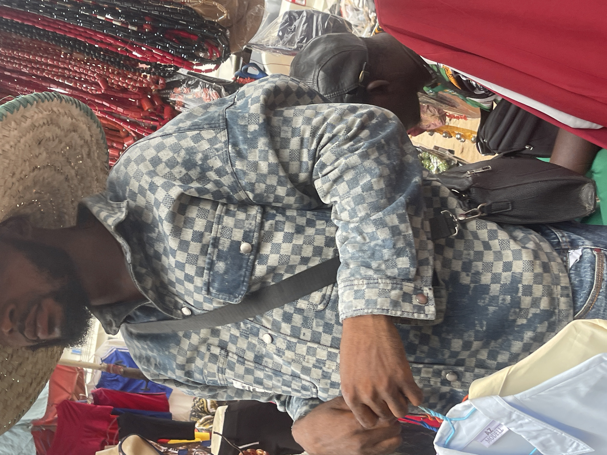 The image portrays a bustling market scene, consistent with the provided location context of Onitsha, Nigeria. In the foreground, a person is depicted from a high angle, looking downwards and engaged with items. This individual wears a wide-brimmed straw hat, possibly with subtle glitter, and a distinctive blue and light grey checkered jacket, which appears to be made of denim or a similar material. A black shoulder bag with a silver buckle is visible, along with a black strap worn across their chest. Their right hand is holding or adjusting several colorful garments on hangers, suggesting activity as either a vendor or a customer.
Further into the background, the head and shoulder of another person wearing a black cap can be seen. The surrounding environment is densely packed with market goods. To the upper left, numerous strands of red and black beaded necklaces are displayed, possibly hanging for sale. Various articles of clothing, including red, blue, cream, and white shirts, are visible on hangers. Notably, a white shirt bears a visible label with the text "AZ MODELL." Other elements in the background include black and green bags and items partially obscured by plastic wrapping, all contributing to a vibrant and crowded commercial atmosphere. The lighting suggests daytime, likely within a covered market area.