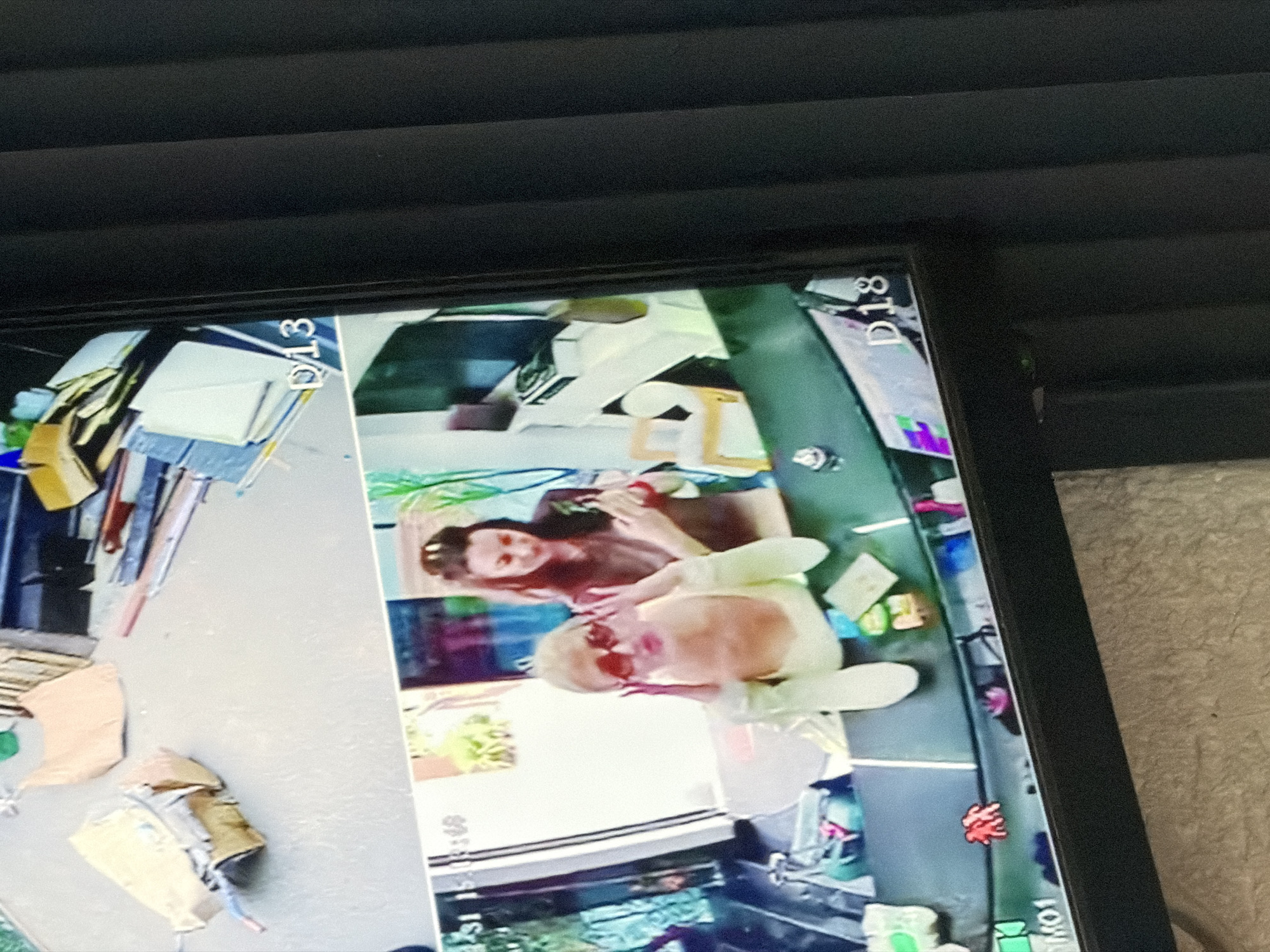 A monitor displays two video feeds, labeled D13 and D18. D13 shows an exterior space with construction materials. D18 depicts two individuals, one wearing sunglasses and a yellow garment. The location is unknown and there is no forensic data to indicate the city of Küçükçekmece, Turkey.