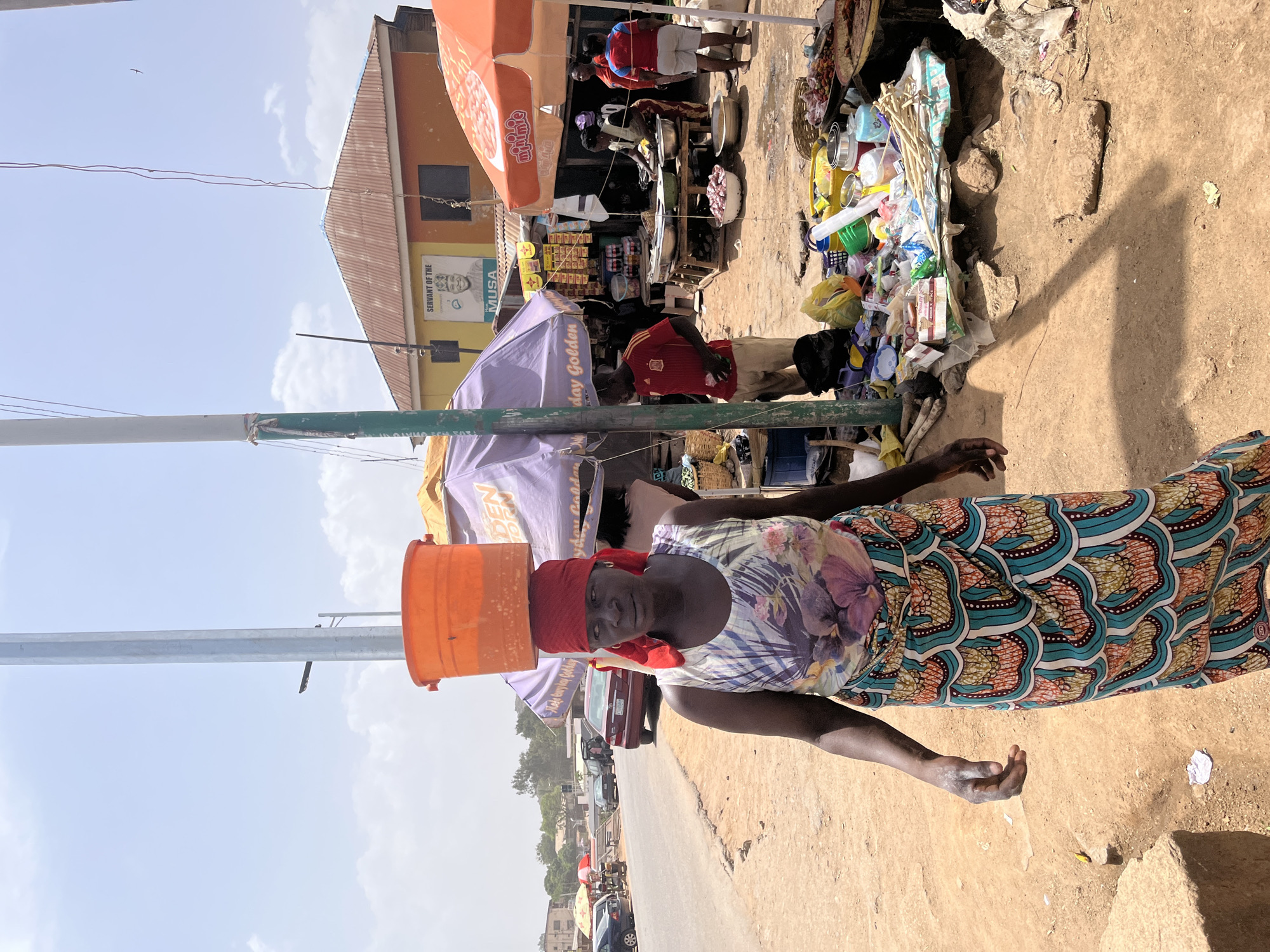 A woman carries a bucket on her head in Gwong, Nigeria. The woman, dressed in a floral top and patterned skirt, balances an orange bucket on her head. A red cloth is tied around her head. She stands on a dirt road alongside a busy street. The background includes market stalls under colorful umbrellas, pedestrians, and vehicles. Buildings are visible beyond the stalls.