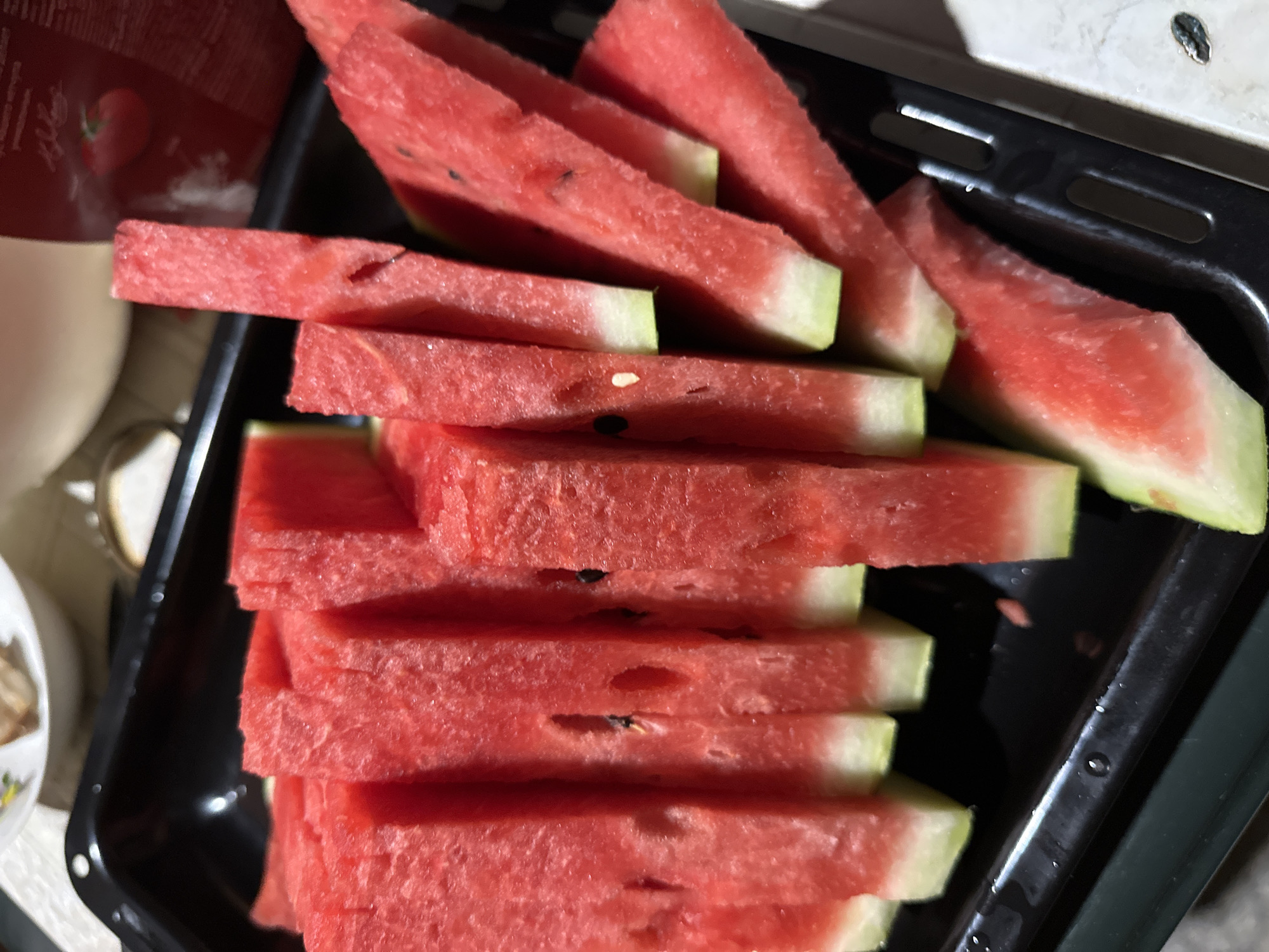 A close-up shot shows a black rectangular baking dish filled with cut watermelon slices. The watermelon is bright red with a thin white rind and a lighter green outer rind, arranged in a dense, slightly overlapping manner. Some slices display black seeds embedded within the red flesh. The dish itself appears to have a slightly textured surface and some moisture droplets. In the background, a blurred white plate with what appears to be food is visible, along with a red object, possibly packaging with text, also out of focus. The scene suggests a domestic setting, likely a kitchen or dining area. No individuals are present in the frame.
