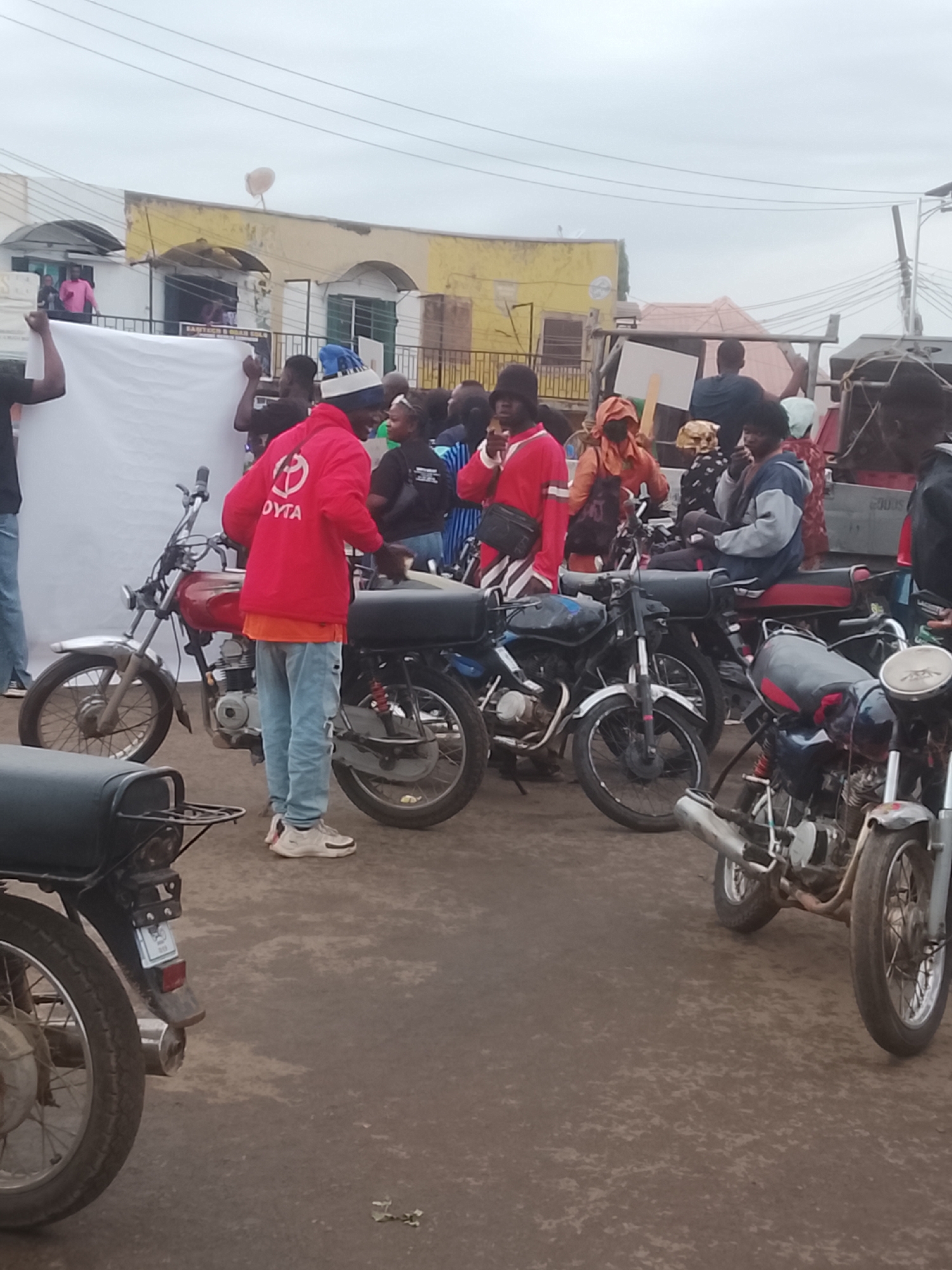 The image depicts a gathering of people and motorcycles in what appears to be a street in Gwong, Nigeria. Several motorcycles are parked closely together. A group of people surround them, with some individuals standing near a large white sheet or banner. One person wears a red jacket with the letters "DYTA" on it. Others are dressed in casual clothing. The buildings in the background are a mix of yellow and white, with a satellite dish visible on the roof. The sky is overcast. Some people are on top of the vehicles.