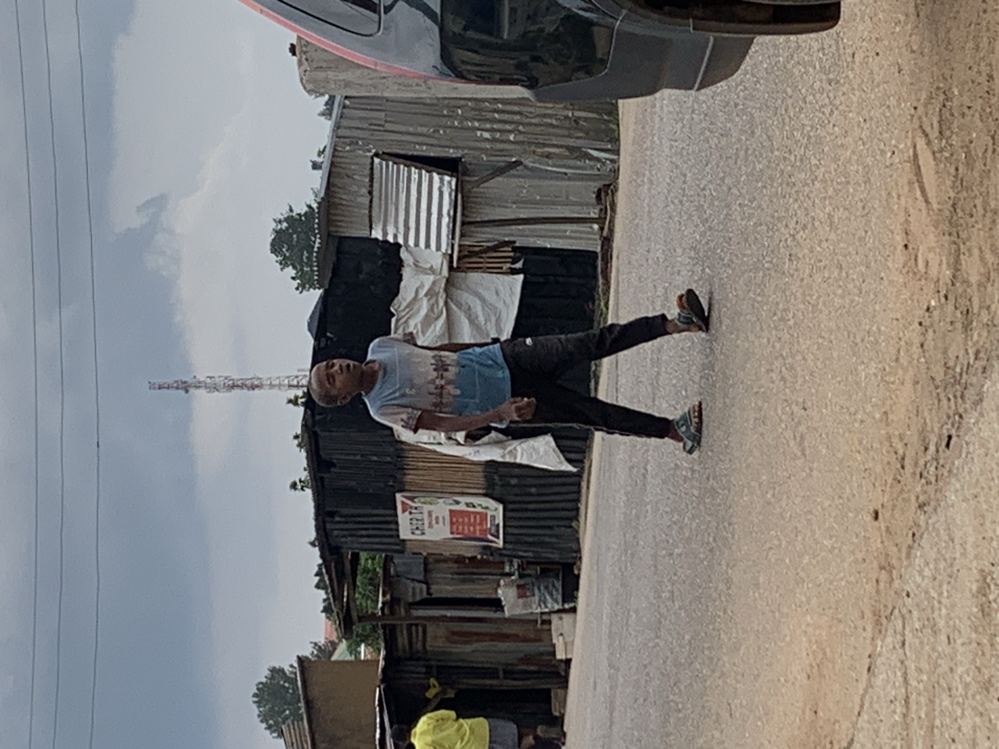 A young boy walks down a dusty, paved road in Jos, Nigeria. He is wearing a blue and white t-shirt with a city skyline graphic, and black athletic pants. He has on sandals. He is walking towards the viewer and looking to the right. The boy is in the middle of the road, with a black car partially visible on the right side of the frame. On the left side of the road are several structures made of corrugated metal, appearing to be makeshift shops or homes. A tall, thin communication tower is visible in the background, against a cloudy sky. The overall environment appears to be an urban or peri-urban area with informal settlements.