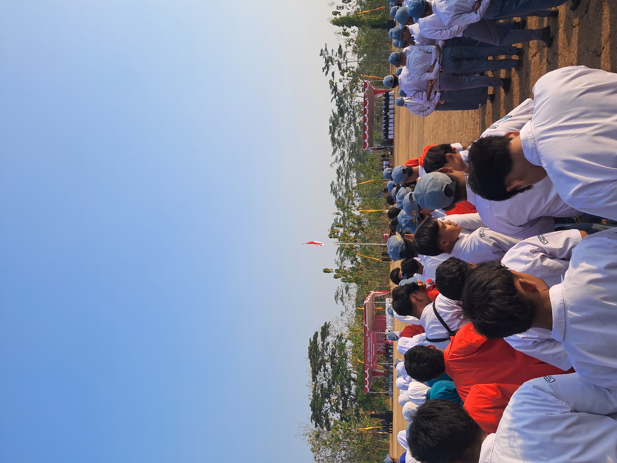 The image depicts an outdoor gathering, likely a school event, taking place in the city of Ponjong, Indonesia. A large group of students, primarily in school uniforms consisting of white shirts and grey or blue caps, are assembled on a field. Some students are wearing red shirts. In the background, a stage is set up with a red and white striped awning, and a banner that reads "UPACARA PERINGATAN" is visible, suggesting a commemorative ceremony. A flag pole with the Indonesian flag is also present. The environment is characterized by a clear blue sky and scattered trees in the background, indicating a dry, possibly sunny, day. The students appear to be standing in formation, facing the stage, and some are looking down or talking amongst themselves, indicating a paused moment in an event. The overall scene suggests an organized event taking place in an open, communal space.