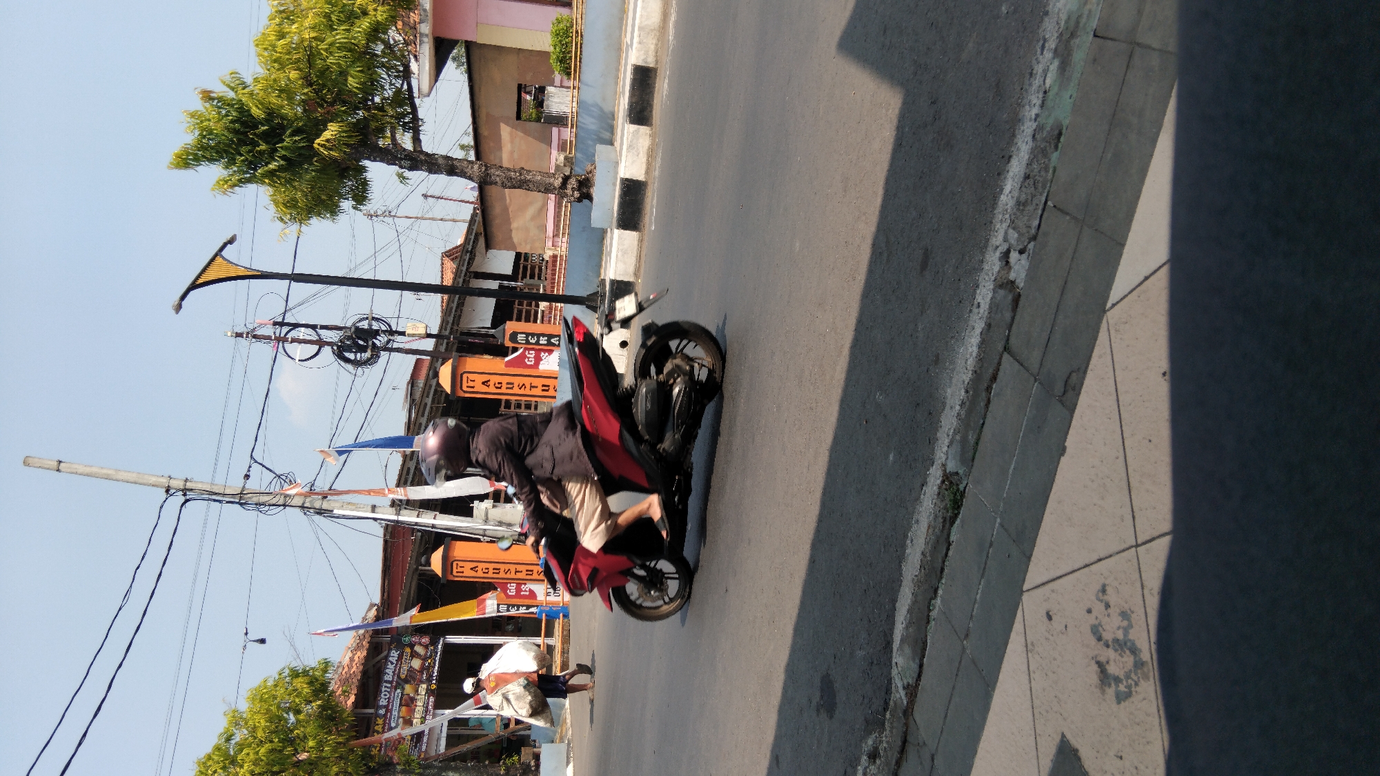 A person wearing a dark jacket and light-colored shorts rides a red scooter on a street in Batang, Indonesia. The rider wears a helmet. The scooter is moving from left to right in the frame. Behind the scooter, there are buildings with decorative pillars. Flags with text that appears to be in Indonesian are visible. A street lamp with a yellow top is also present. To the left of the scooter, another person is walking away from the camera, carrying a large white bag. The street has a concrete curb and sidewalk. The sky is clear and blue. The image is taken outdoors during the day.