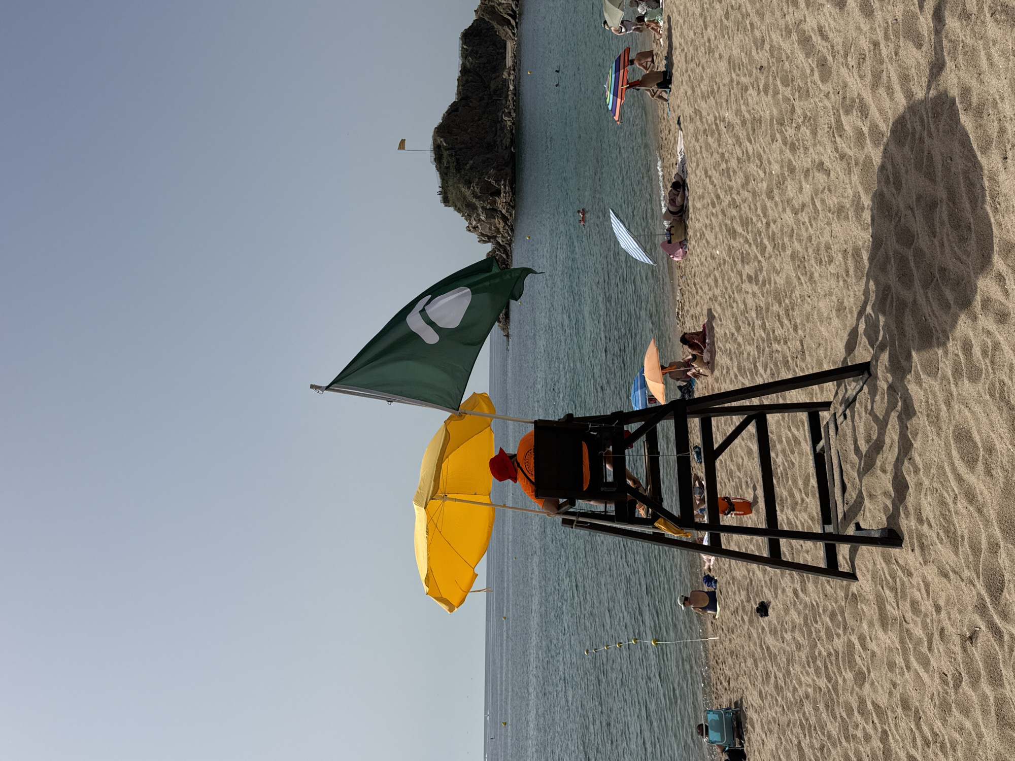 A lifeguard is seated on a tall, dark wooden chair on a sandy beach in Blanes, Spain. The lifeguard wears an orange shirt and a red hat and is shaded by a bright yellow umbrella. A large green flag with a white symbol is unfurled to the right of the lifeguard's chair. The sea is calm and blue, with a rocky island in the distance topped with a flag. Several beach umbrellas and sunbathers are visible further down the beach. The sky is clear and bright.
