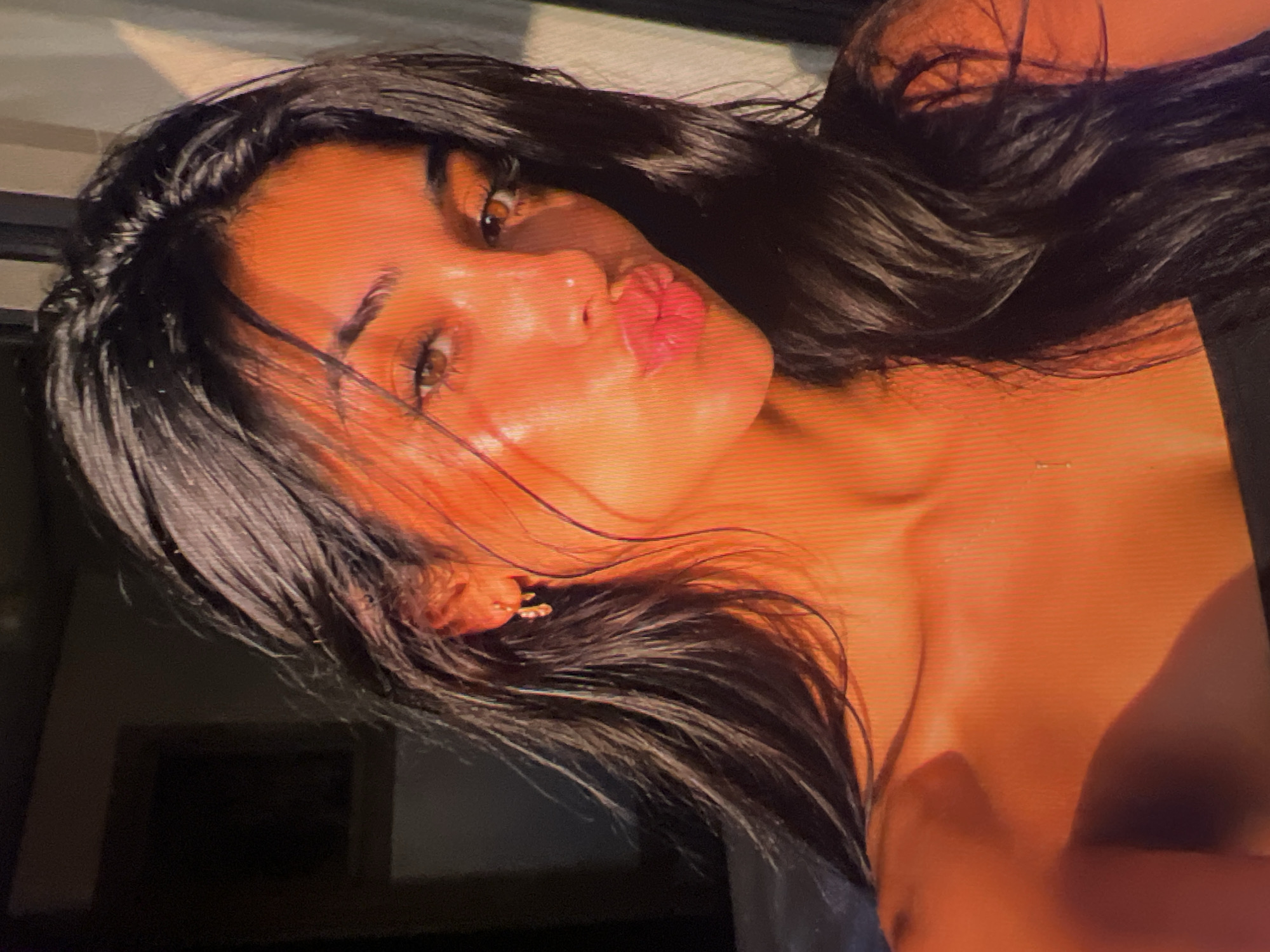 The image shows a close-up, head-and-shoulders shot of a young woman with long, dark hair. She is looking directly at the camera with her lips pursed, as if about to kiss. The lighting is warm and golden, suggesting it might be sunset or sunrise. There is a faint shadow on the left side of her face, and a bright light source on the right, casting a subtle highlight on her skin and hair. She is wearing a dark-colored garment, possibly a top or dress, that exposes her shoulders. A small, delicate earring is visible in her left ear. The background is dark and indistinct, with a hint of a framed picture or object on the far left. The overall impression is one of intimacy and natural beauty. There is no discernible location in the city of Athens, Greece.