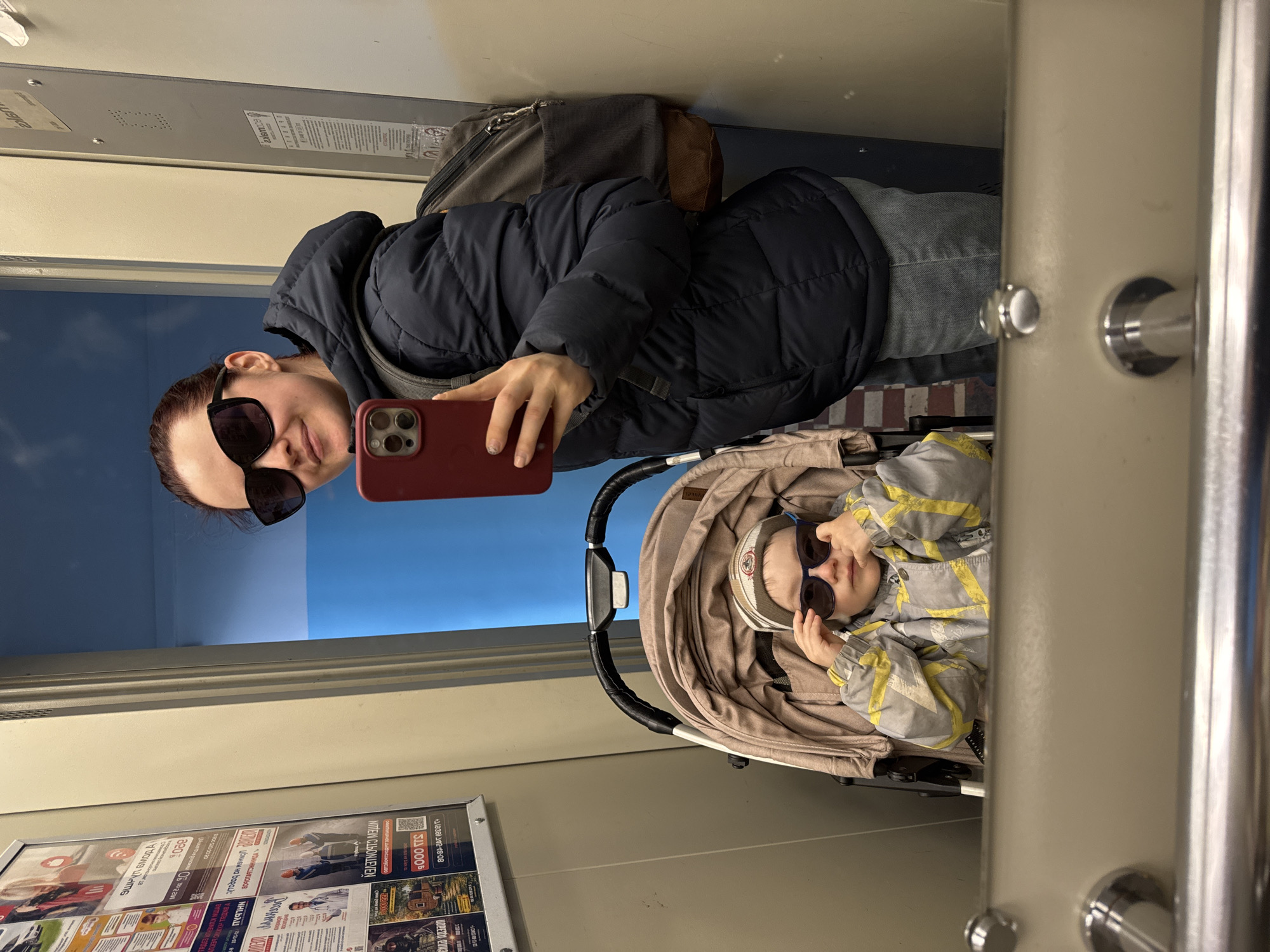 This image is a selfie taken through a reflective surface, likely a window or mirror, inside what appears to be a public transport vehicle in Izhevsk, Russia.
The primary subjects are a woman and a young child. The woman, positioned on the left, has dark hair, wears sunglasses, a dark puffer jacket, blue jeans, and a dark backpack. She holds a red smartphone (resembling an iPhone) to take the picture, showing a slight smile. Next to her, to the right, a young child is seated in a beige stroller. The child also wears sunglasses, a grey jacket with yellow patterns, and a striped hat, looking calmly towards the camera.
The setting is characterized by interior elements of a vehicle, including silver metal poles or handles on the right side of the image, and a ceiling with a ventilation grille. Through the reflection, a bright blue sky is visible, suggesting daytime.
A notable feature of the scene is an advertisement board on the bottom left, displaying various ads with Cyrillic text. Visible phrases include "3 000 руб" (3,000 rubles), a QR code, and a phone number "+7 (912) 420-48-08". Other fragmented Cyrillic words and numbers are present, indicating job postings or service advertisements. The brand "TUCI" is visible on the child's stroller.
The activity taking place is a casual travel moment, with the woman capturing a shared experience with the child. The overall mood conveyed is relaxed.