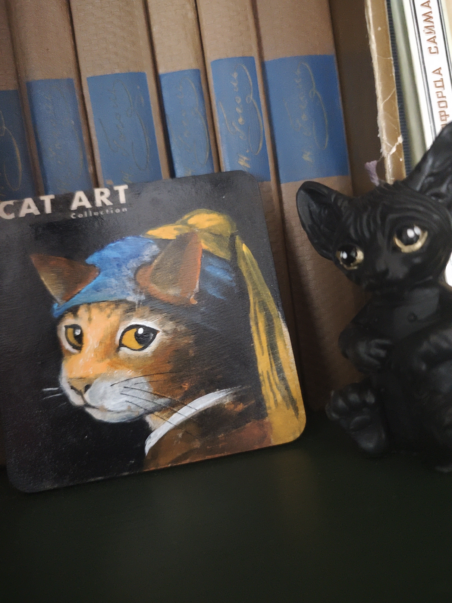 A close-up shot depicts a "CAT ART Collection" item featuring a cat portrait inspired by "Girl with a Pearl Earring", positioned next to a small, glossy black cat figurine. These objects sit in front of a row of books with brown covers and blue spines. The background is slightly blurred, providing context to the subjects. No people are visible. The scene appears to be indoors, possibly on a shelf or table. There is no indication of the location of Свистуха (кузяевский С/о), Russia within the image.