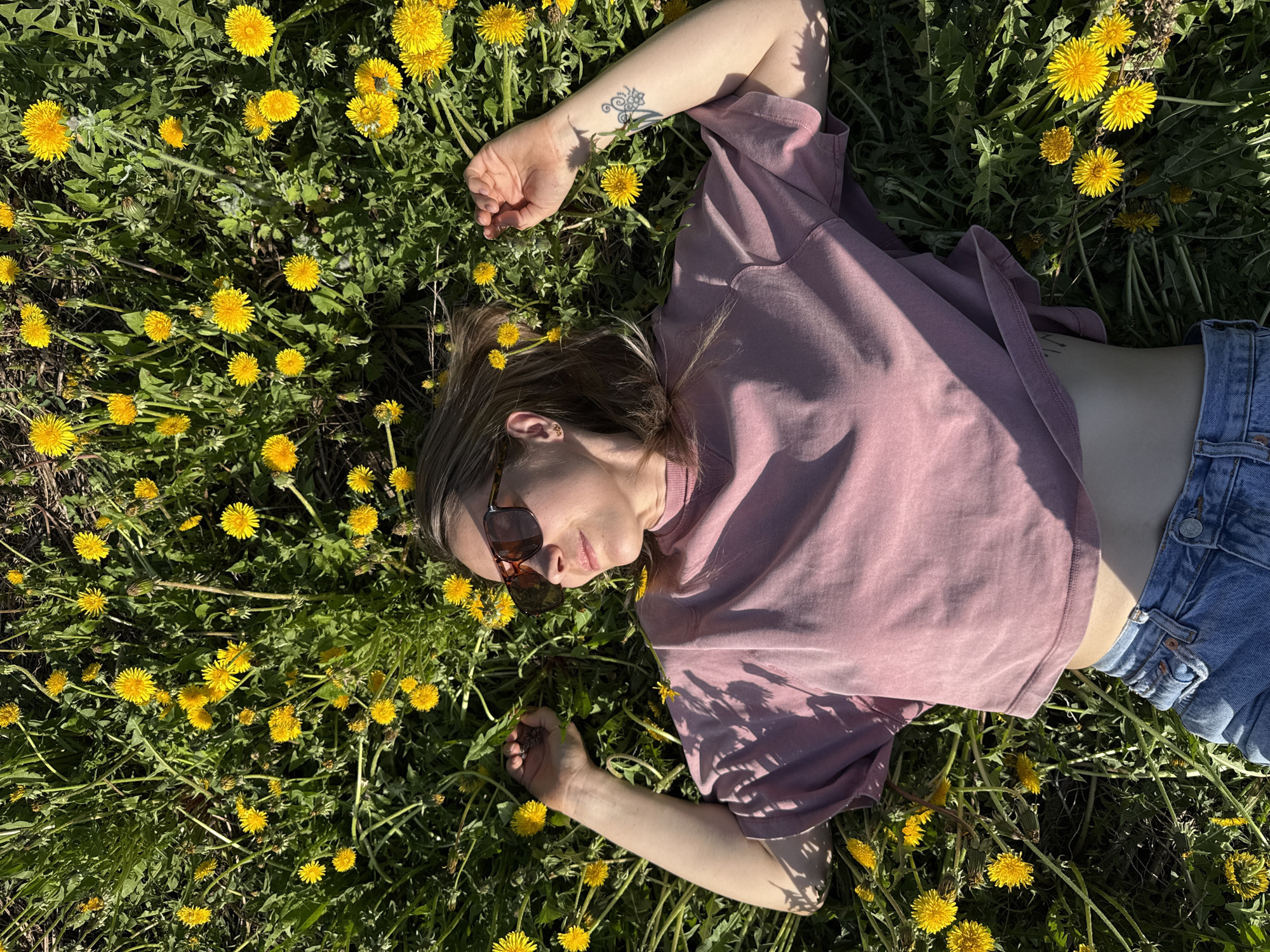 A woman lies in a field of dandelions. She wears a pink t-shirt, blue denim shorts, and sunglasses. She has a tattoo on her left forearm. The location is the city of Chaikovsky, Russia.