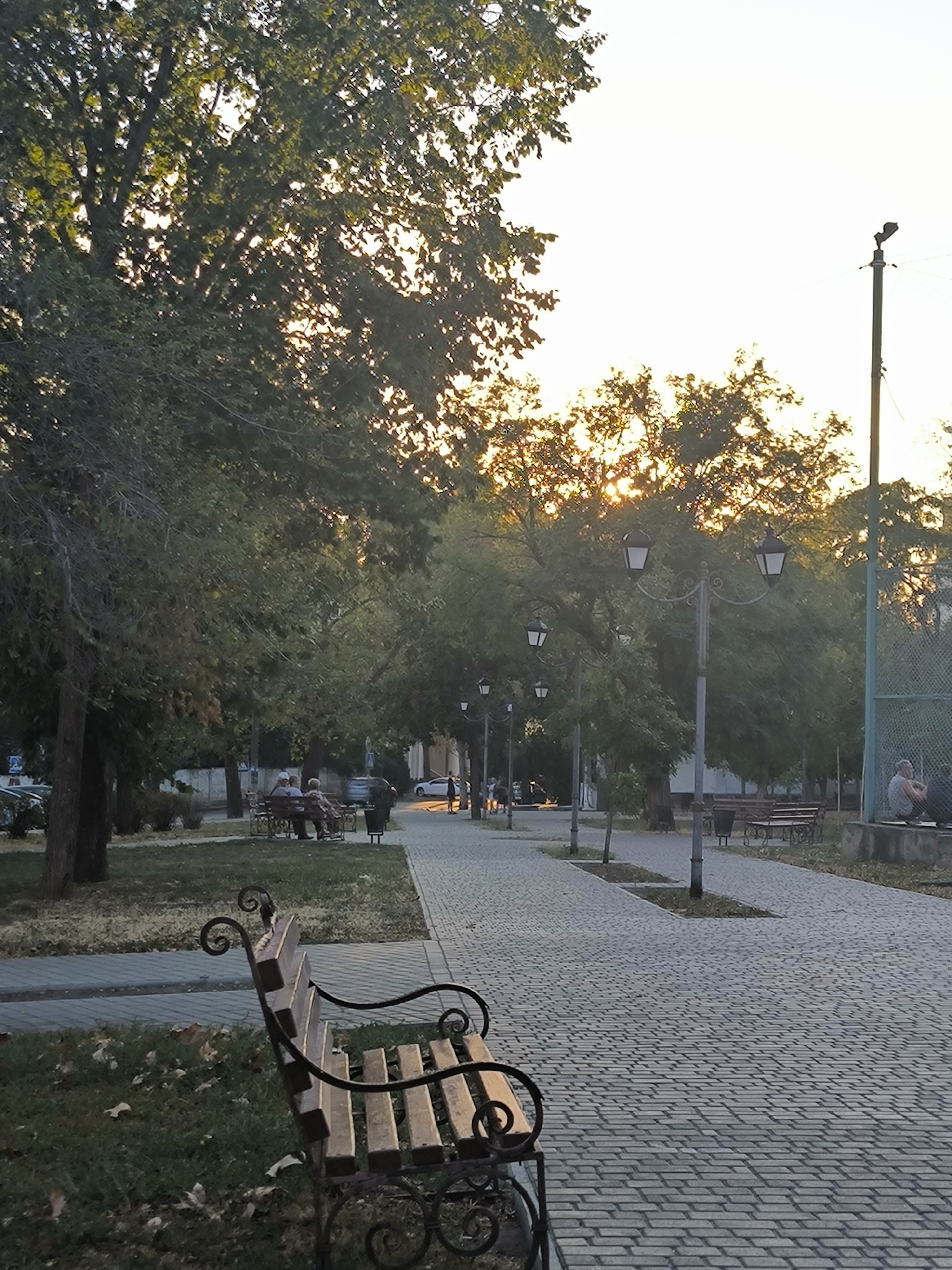 A wide shot captures a tree-lined parkway in Mykolaiv, Ukraine, bathed in the warm glow of setting sun. The paving stones of the walkway are a mosaic of grey, leading the eye towards a cluster of trees with dappled sunlight filtering through their leaves. On the left, a bench with ornate metalwork and wooden slats sits in the foreground, facing a patch of grass dotted with fallen leaves. In the middle ground, two individuals are seated on another park bench, appearing to converse. Further down the path, more benches and lampposts are visible. To the right, a chain-link fence encloses a sports court, where a single individual sits on a ledge. The sky above is a pale, washed-out blue, with the sun's rays creating a bright haze.