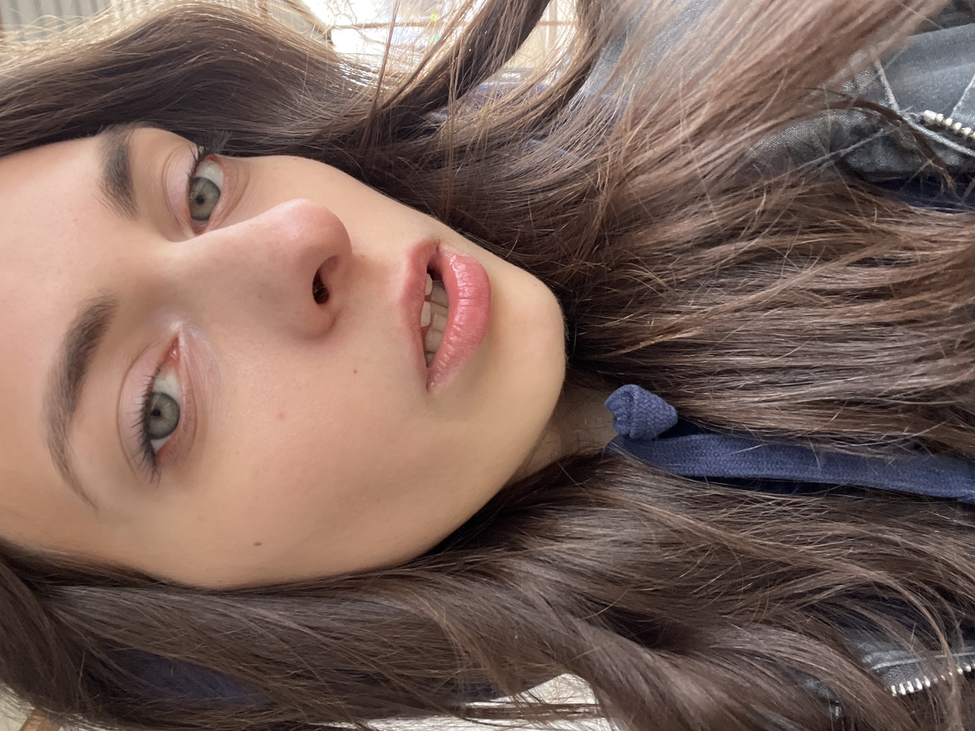 A close-up selfie of a young woman with long, wavy brown hair. Her face is turned slightly to the side, with her eyes looking towards the camera. She has light skin, full lips, and green eyes. She is wearing a blue hoodie and a jacket with a zipper. The background is blurry and out of focus, but appears to be indoors. There is no way to definitively determine the location to be in Krasnoyarsk, Russia based on this image alone.