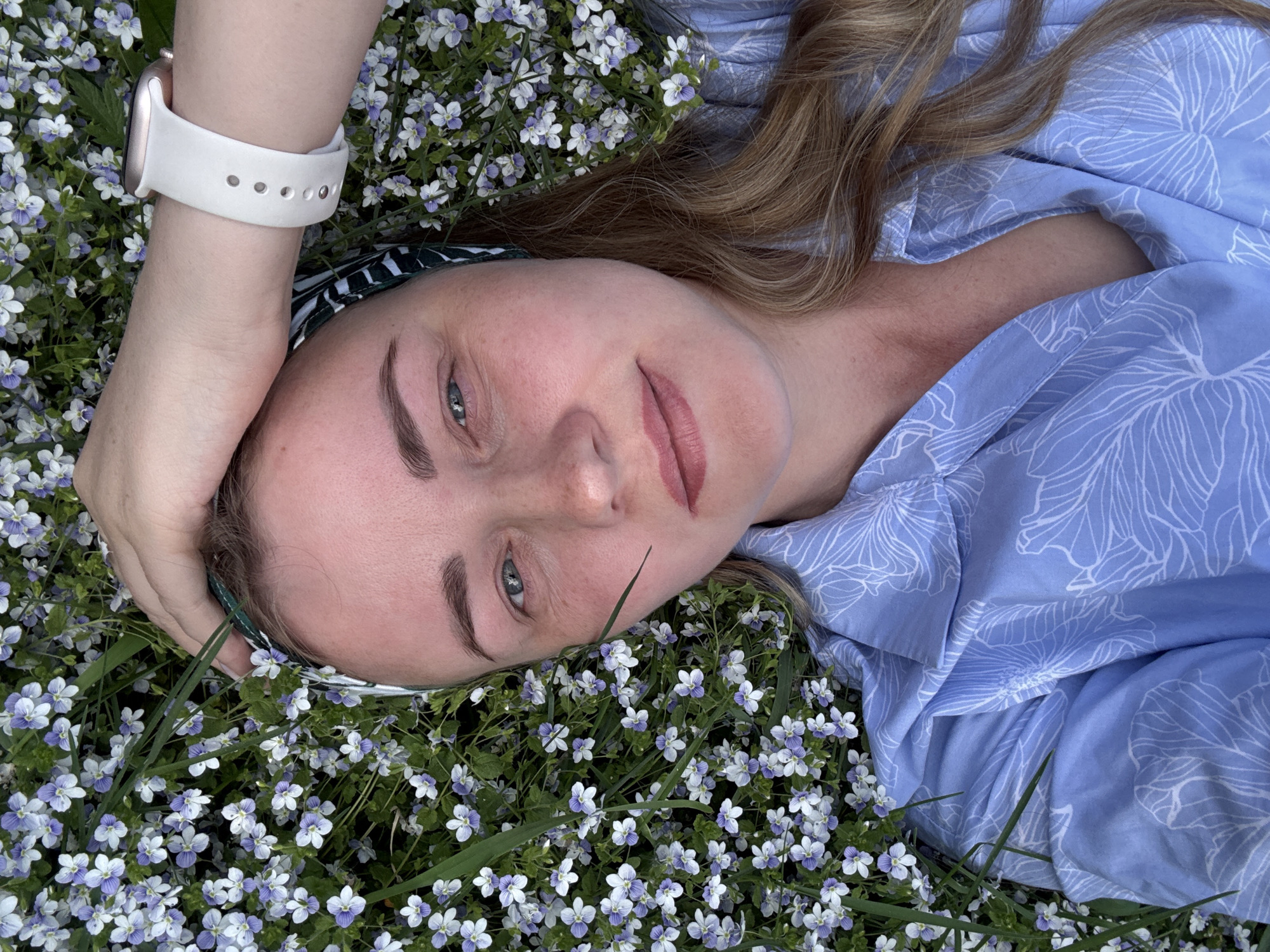A woman lies on her back in a field of small, white and purple flowers. She is wearing a light blue patterned shirt, a headband, and a white smartwatch. Her hair is blonde and partially covers her right shoulder. Her right arm is bent and her hand is resting on her forehead. The background is filled with the flowers. The location appears to be Chaikovsky, Russia, based on prior knowledge, although this is not immediately clear from the image.