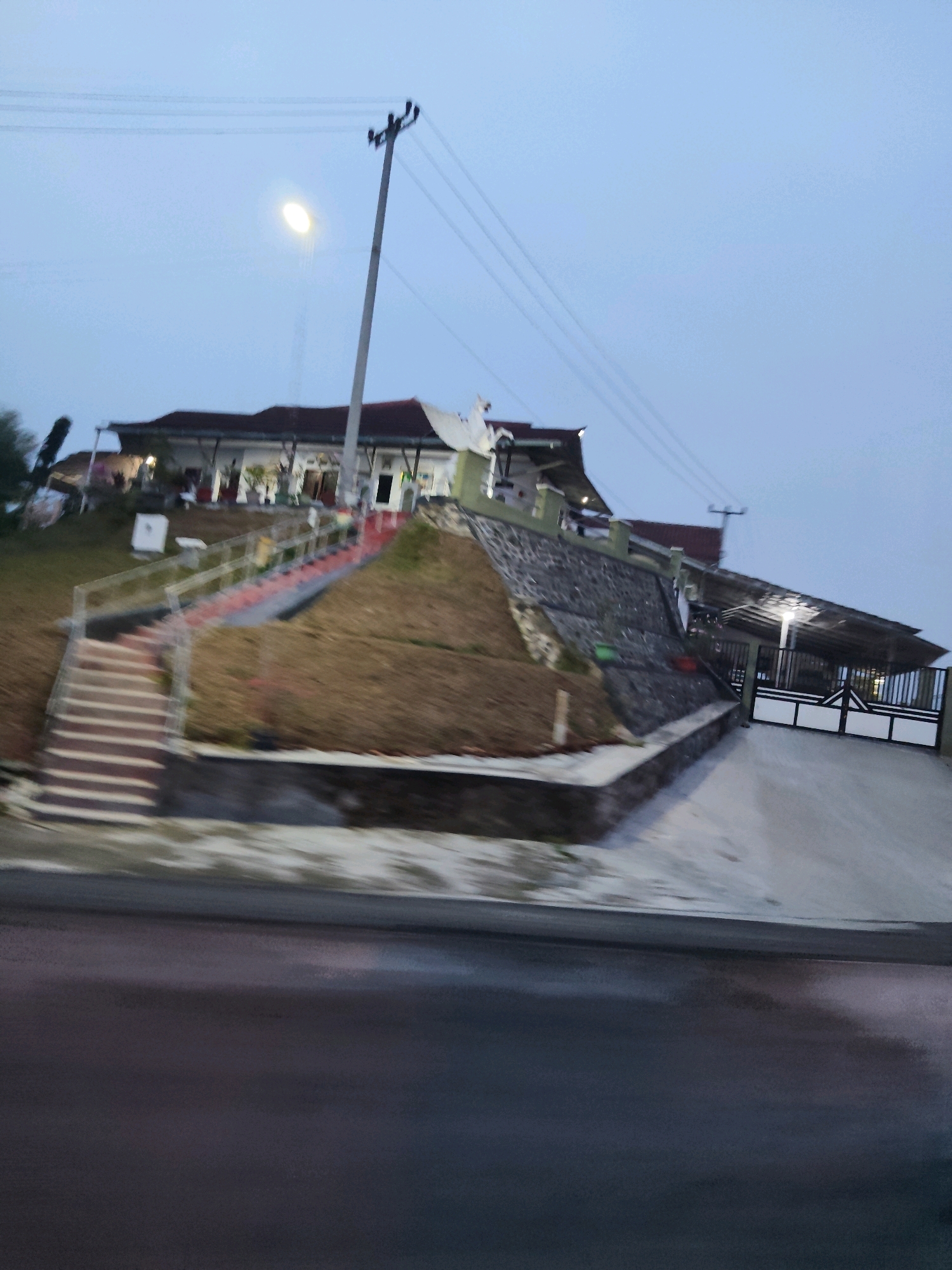 The image depicts a building situated on a slope, accessed by a long, red-lined staircase and a paved driveway. The building appears to be a residential property or perhaps a small establishment, with a covered patio area and a large white and black gate. The environment is overcast and possibly humid, with some vegetation visible on the hillside. A utility pole with electrical lines stands prominently to the right of the building. The road in the foreground is wet and reflective, suggesting recent rain or damp conditions. The overall impression is that of a slightly blurry, twilight or early morning scene in a suburban or semi-rural area.