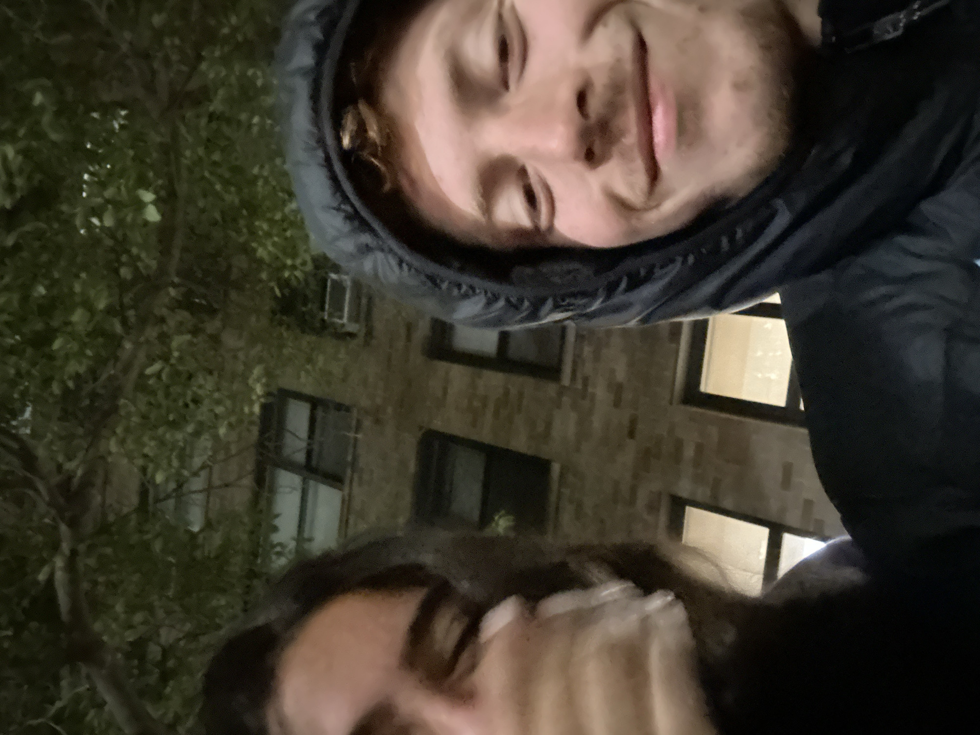 The image is a rotated, low-angle selfie featuring two individuals, likely taken at night or dusk in an urban environment.
The upper portion of the frame is dominated by a fair-skinned male subject, seen from the chin up. He has reddish-brown hair partially visible under a dark, hooded jacket. He has light stubble on his face and a slight, pleasant smile. His gaze is directed towards the camera.
Below him, only a portion of a second individual's face, dark hair, and shoulders are visible. This person appears to be wearing a dark garment and is also looking towards the camera, but their expression is less discernible.
In the background, behind the subjects, are dark green trees with visible foliage, suggesting an outdoor setting. A multi-story brick building is also visible, with several windows that are illuminated from within, indicating artificial light and reinforcing the night/dusk time of day. The building's architecture, combined with the trees, suggests a street or park area in a city, consistent with a New York location.
The overall activity is a casual photograph or selfie. There is no visible text in the image.