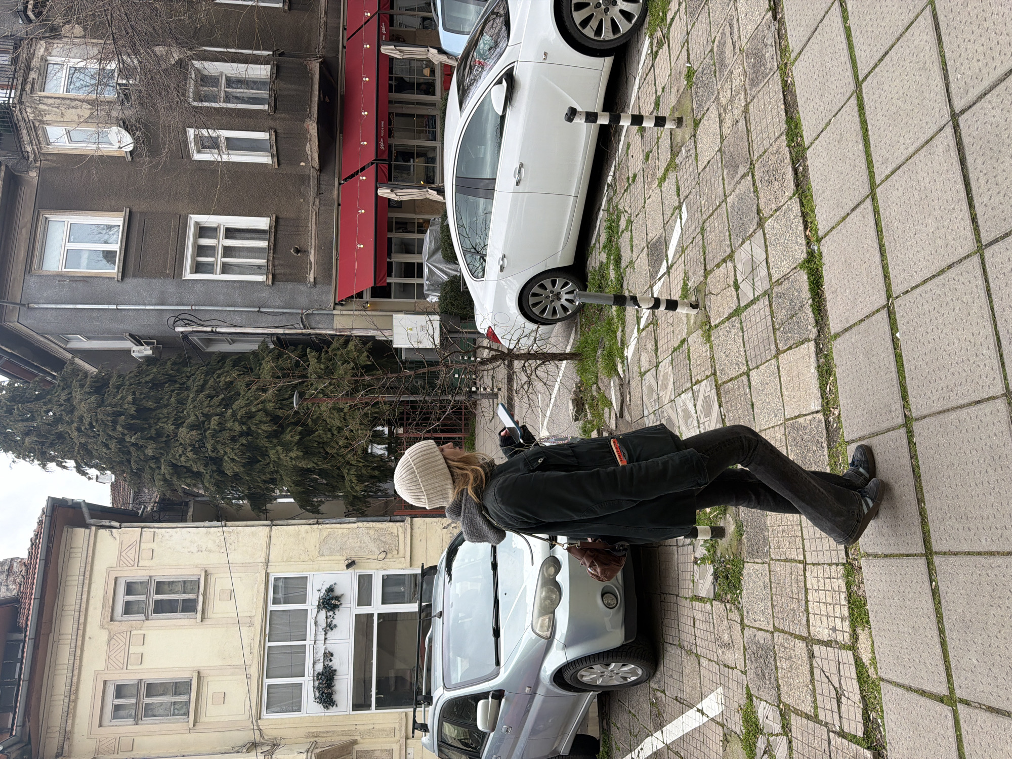 The photo, taken in Sofia, Bulgaria, shows a young woman standing on a sidewalk. She is wearing a white knit cap, a gray scarf, a black jacket, dark jeans, and dark shoes. She holds a phone in her left hand and a brown purse hangs from her shoulder. She is standing between a silver SUV and a white car parked along the sidewalk, positioned between black and white striped bollards. The sidewalk is made of square concrete paving stones with grass growing in the gaps. Behind her are buildings with architectural details; one is a light brown color, and the other is gray with a red awning. A large evergreen tree is visible between the buildings. The sky is overcast.