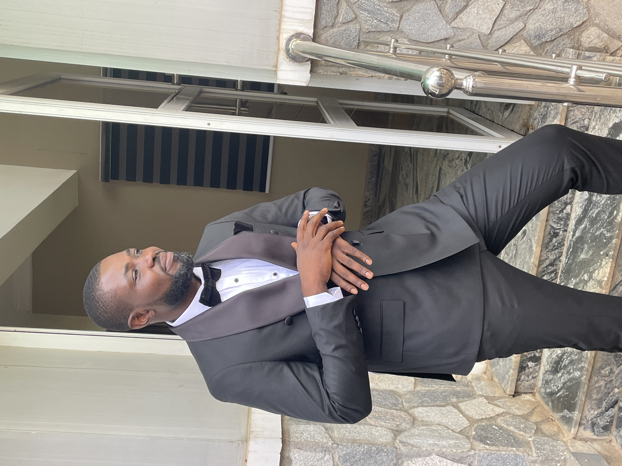 A man wearing a black tuxedo with a bow tie is standing outside a building. He has short hair and a beard. His hands are clasped together in front of him. The building behind him has a stone facade. There are metal railings visible. The exact location in Bukuru, Nigeria cannot be determined from the image.