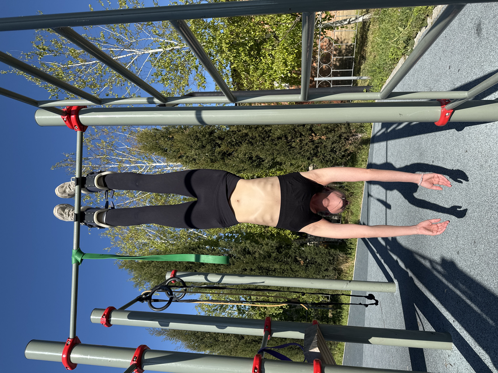 A woman in a black crop top and black leggings is hanging upside down from a metal playground structure in городское поселение Щелкино, Ukraine. Her feet are secured in gray shoes with straps attached to the structure. Her arms are extended downward. The playground has gray metal bars with red connectors. Trees, grass, and a blue sky are visible in the background.