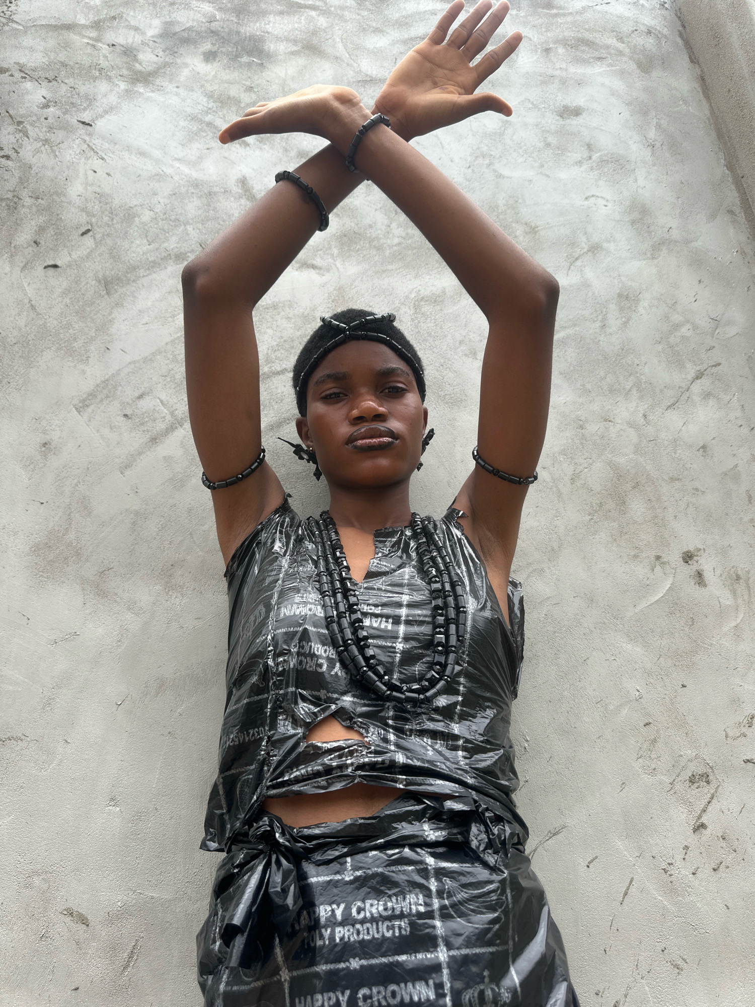 A young person, appearing to be of African descent, stands against a rough, grey concrete wall in what is described as Badagry, Nigeria. They are positioned centrally, facing the camera, with their arms raised and crossed above their head. Their attire is unconventional, consisting of what appears to be black plastic material, possibly packaging, fashioned into a top and a skirt. The material is textured with a repeating grid pattern and text, some of which is visible and reads "HAPPY CROWN TOLY PRODUCTS". There are rips and tears in the plastic fabric, revealing the person's midriff and skin. They are adorned with multiple black beaded necklaces and bracelets, as well as a black beaded headband. The lighting suggests an outdoor setting, with natural light illuminating the scene from above. The background is a textured, weathered concrete surface, showing signs of age and wear. The composition is a waist-up shot, with the focus on the individual and their unique presentation.