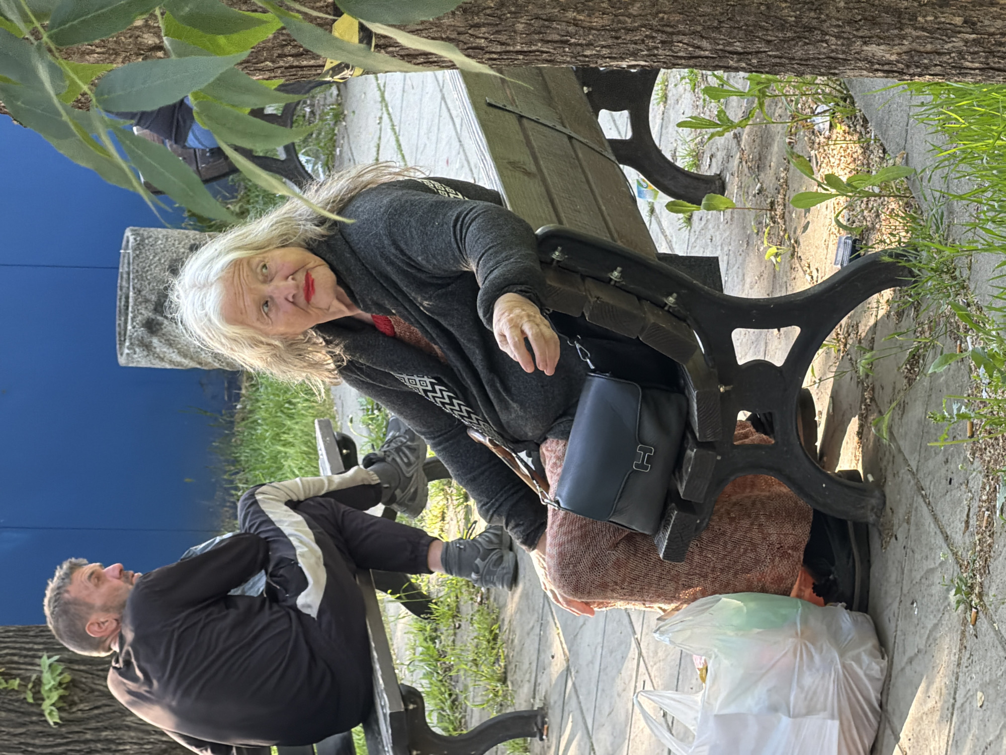 A woman with white hair sits on a park bench beside a man. The woman wears a dark gray cardigan over a reddish-brown garment. A dark blue purse hangs from her left shoulder. She looks toward the camera. A man reclines on the bench beside her. He wears a black tracksuit with white stripes. Plastic bags are at the woman's feet. The scene is outdoors, adjacent to a tree and other vegetation, likely in a park in Sofia, Bulgaria.