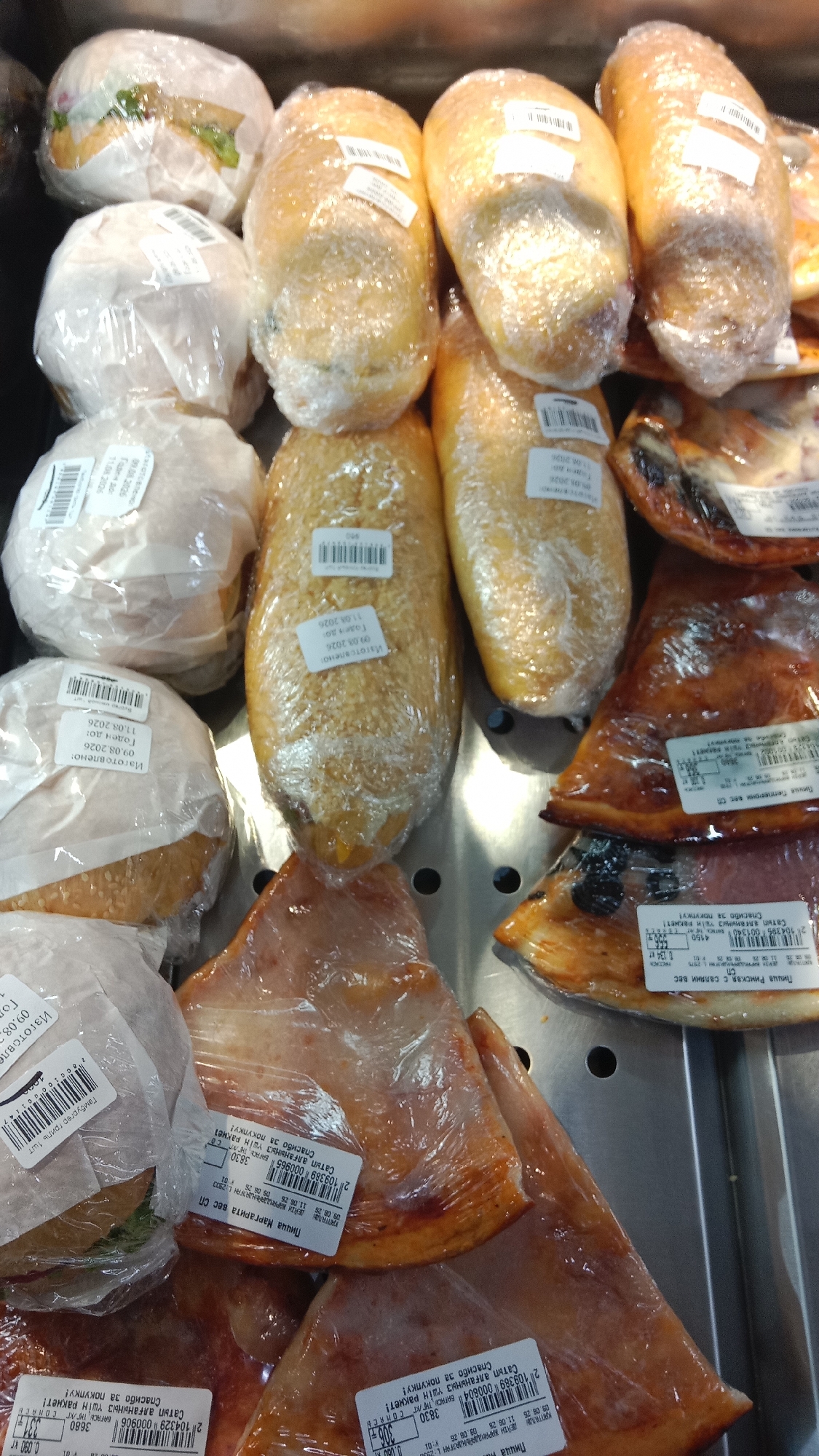 The image shows a display of food items, primarily bread products and what appear to be meat products, all individually wrapped in clear plastic. The items are arranged in rows on a metallic shelf, suggesting a retail environment such as a grocery store or market. Several of the items have white labels with black text, likely indicating product information, prices, or expiration dates. Some labels appear to be in Russian. The lighting suggests an indoor setting, possibly illuminated by fluorescent lights, as is common in such establishments. The overall impression is one of a food display ready for sale. Without further context or visible landmarks, it is not possible to determine the specific location within Petropavl, Kazakhstan.
