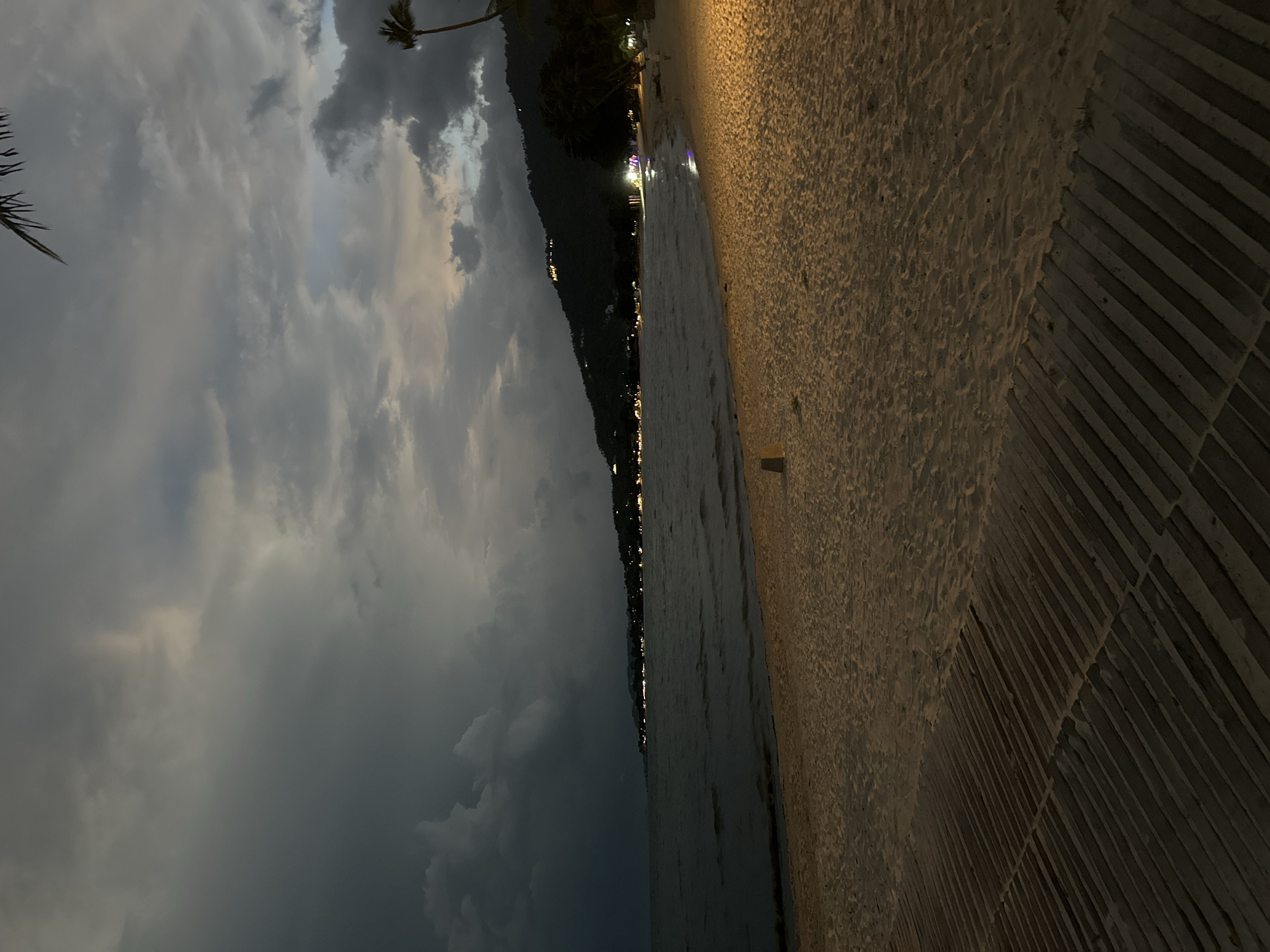 The image depicts a beach in the Ko Samui District of Thailand at dusk or night. The sky is filled with dramatic, dark clouds, suggesting an impending storm or the aftermath of one, with some lighter hues of sunset or artificial light visible on the horizon. The sea is dark and appears calm with gentle waves lapping at the sandy shore. Along the coastline, a line of lights indicates buildings or resorts nestled on a dark, tree-covered hillside. The foreground features a sandy beach with a wooden walkway made of parallel slats. There is a small, light-colored post or marker on the sand. A single palm tree is visible on the right side of the image, silhouetted against the sky. The overall atmosphere is subdued and moody, characteristic of twilight or nighttime on a tropical beach. There are no people clearly visible in the scene.