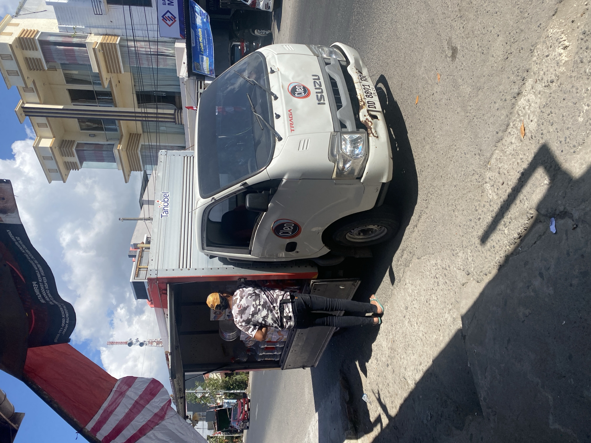 In Bulukumba, Indonesia, a man wearing a camouflage t-shirt, black pants, and a yellow cap stands beside an open-back white Isuzu truck, loading large plastic water containers. The truck has "Tanobel" and "Cleo" branding. The scene takes place on a paved street during the day, with a multi-story building visible in the background, featuring modern architecture with beige and cream colors and multiple windows. A red car is also visible driving on the street. The sky is partly cloudy. The truck's license plate reads "DD 8891 RR".