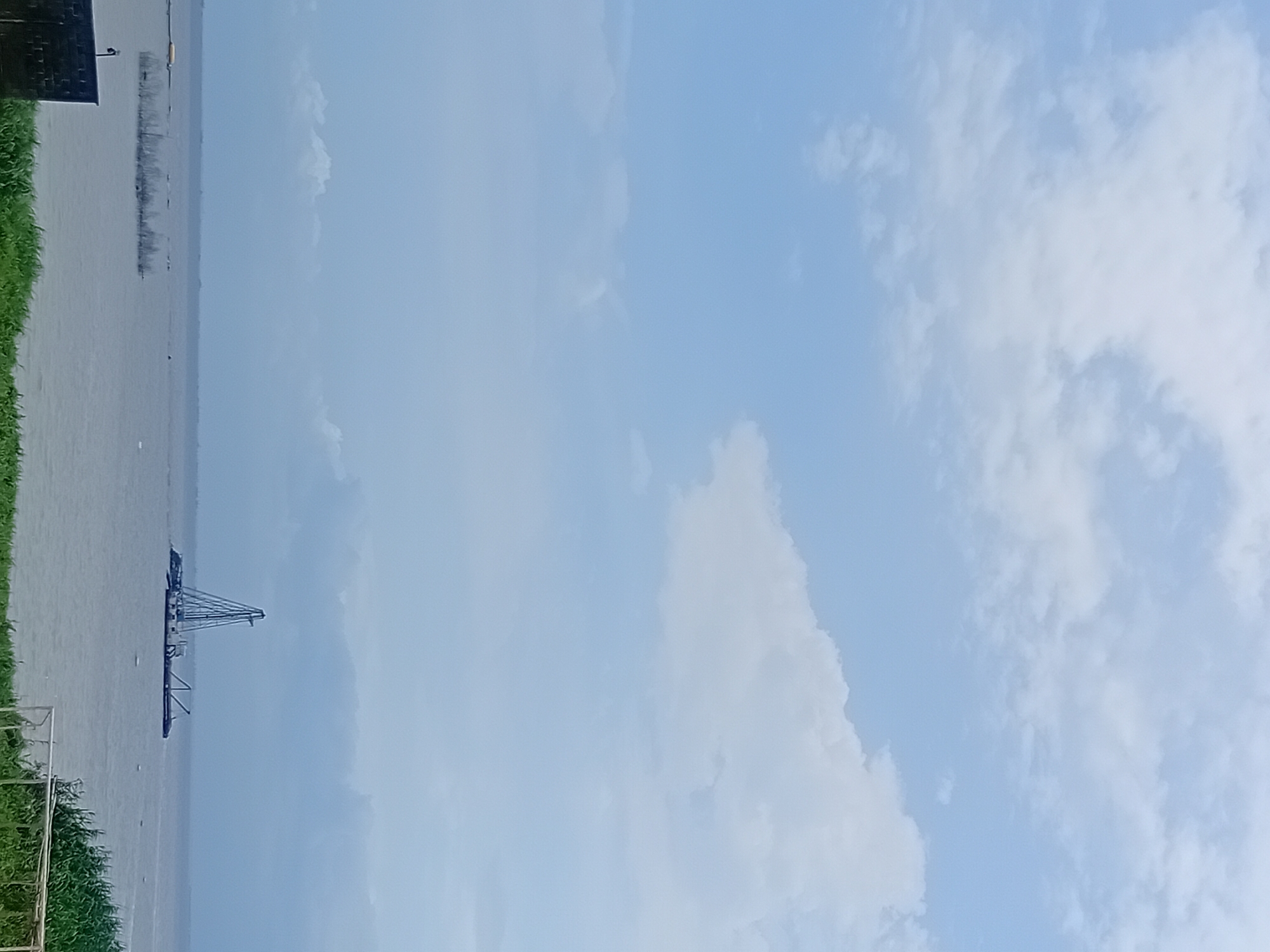 The image shows a wide, calm body of water under a cloudy, light blue sky. In the distance, a large barge with a tall, skeletal tower structure is positioned on the water. The barge appears to be a crane barge. The water is a muted gray-blue color, and a thin line of land is visible on the horizon. In the foreground, there is a patch of green grass and a section of a white metal fence. To the left, a dark, rectangular structure partially enters the frame. The overall atmosphere is overcast and serene.