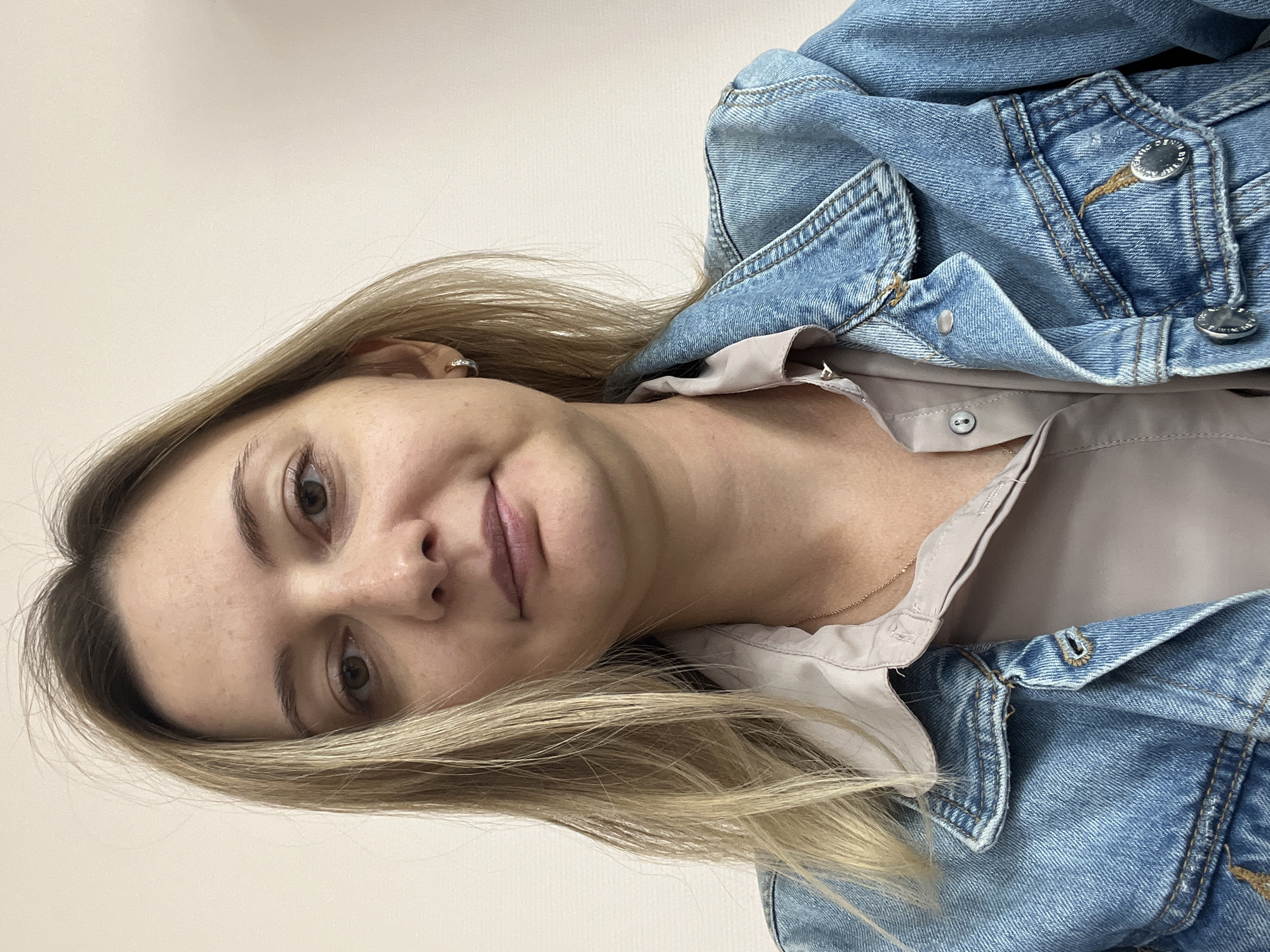 The image shows a medium shot of a blonde woman in a denim jacket and a light-colored shirt. She has fair skin and is looking directly at the camera. She is wearing a small hoop earring in her left ear and a delicate necklace. The background is a plain, light-colored wall, suggesting an indoor setting. There are no visible clues in the image to indicate the location within Volgograd, Russia.